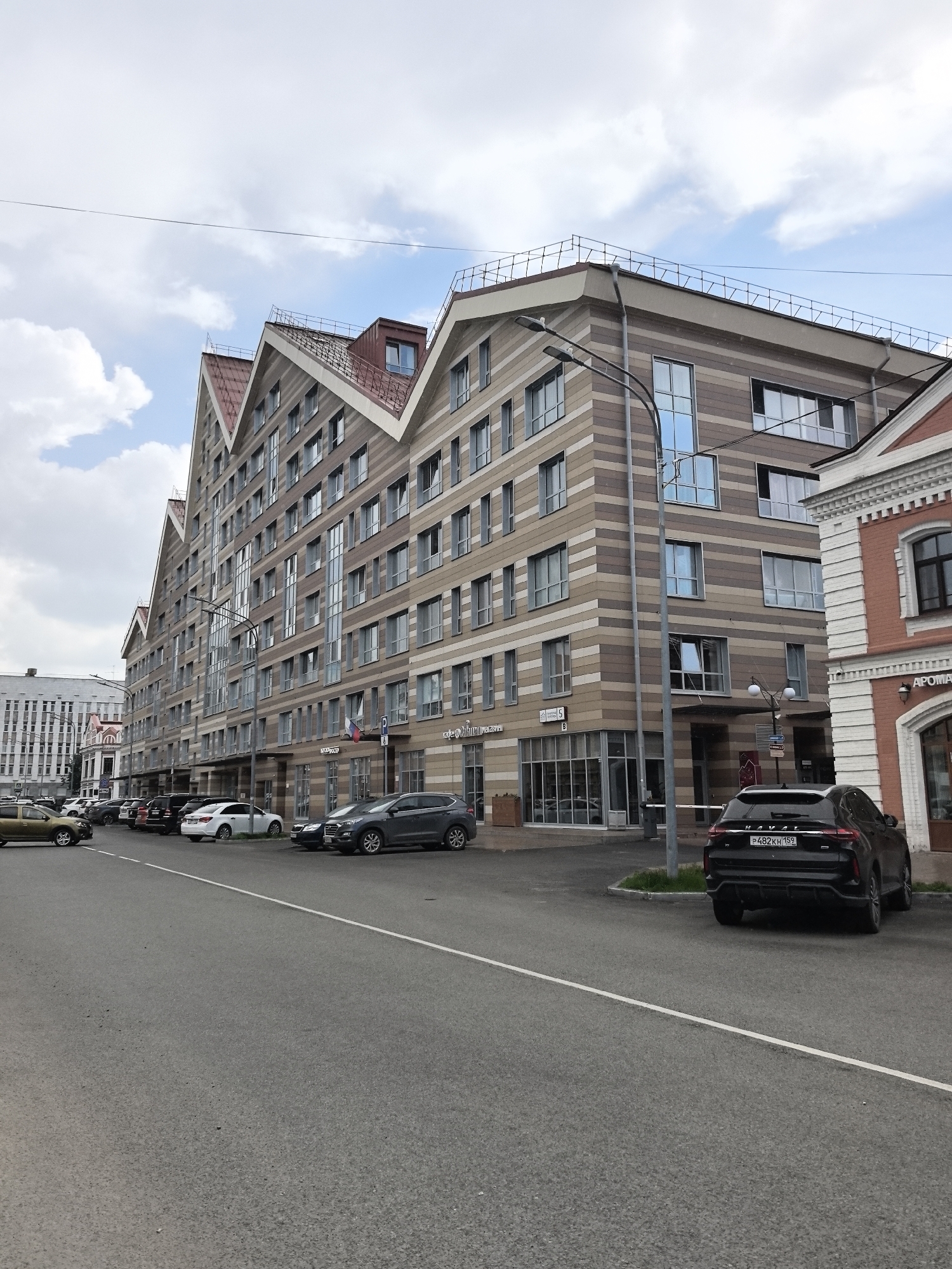 The image shows a street scene in Perm, Russia, featuring a modern, multi-story building with a distinctive, staggered roofline. The building is constructed with alternating bands of light and dark horizontal siding, giving it a textured appearance. Numerous windows are visible across its facade. In front of the building, several cars are parked along the street. A black SUV is prominently positioned in the foreground, facing away from the camera, with its license plate visible. To the left, other vehicles, including sedans and a light-colored SUV, are parked in a row. The street itself is paved asphalt with a white dividing line. A lamppost stands near the building. The sky above is partly cloudy. A smaller, red-brick building with decorative white trim is partially visible to the right of the main building. The overall impression is of a contemporary urban environment.