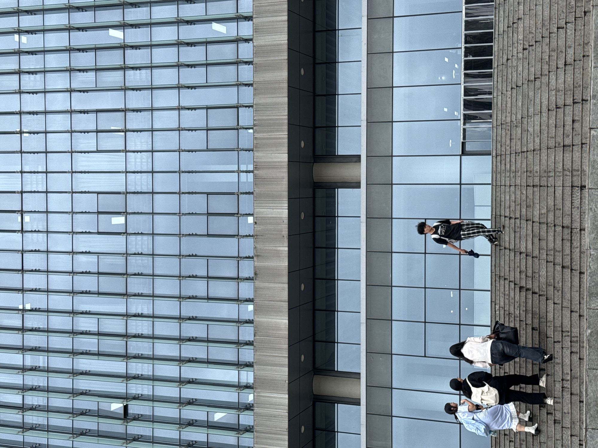 The image depicts a vertical view of a section of an urban landscape, likely in Jiangjin, China, showing pedestrians walking along a grey-tiled sidewalk. To the left, a modern building with reflective glass panels dominates the scene. A vertical grey structure intersects the left and right parts of the building. Four individuals are visible, walking from top to bottom. The person at the top of the frame is dressed in dark casual clothes. Below them, two individuals, one wearing a white top and another wearing dark clothing, followed by a person in a light blue dress. The sidewalk is adjacent to the building.