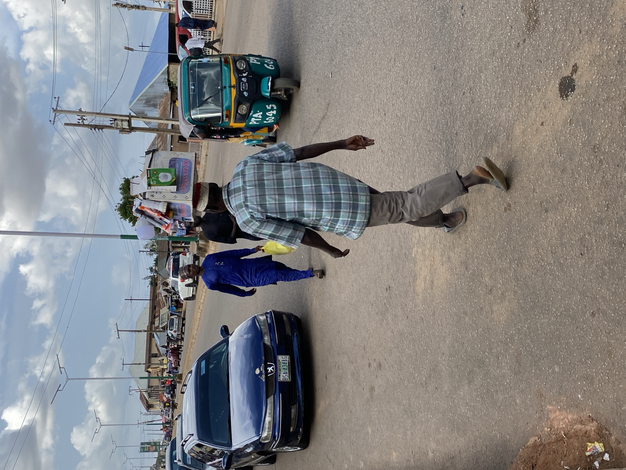 The image captures a street scene in Bukuru, Nigeria. The view is partially obscured and rotated. A blue Peugeot car is parked along the left side of the road. A man in a blue robe walks past the car. Another man in a plaid shirt is walking across the road. A green and yellow tricycle taxi is to the left of the man in plaid. Shops and a line of cars can be seen in the background on the left. The sky is visible above buildings on the left.