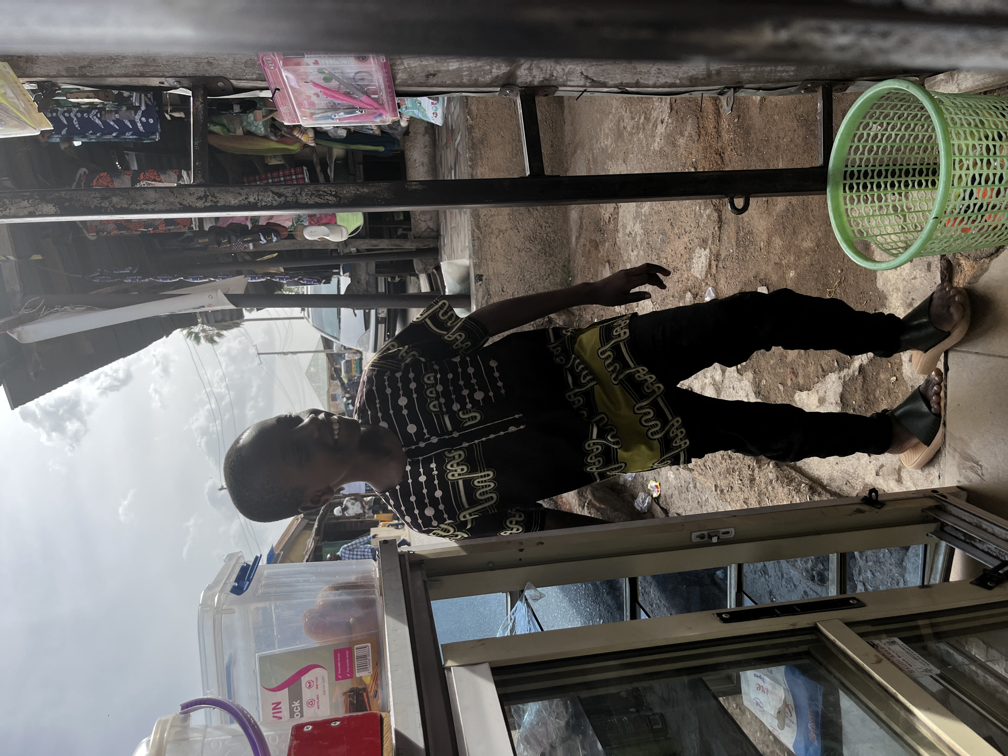 A man stands in what appears to be a small market in Jos, Nigeria. The man is Black, wearing a patterned shirt and black pants. He stands next to a metal frame structure, possibly a stall, with a variety of items displayed, including stationery and containers. A green plastic basket is visible near his feet. The ground is uneven and appears to be made of concrete. The sky is overcast.