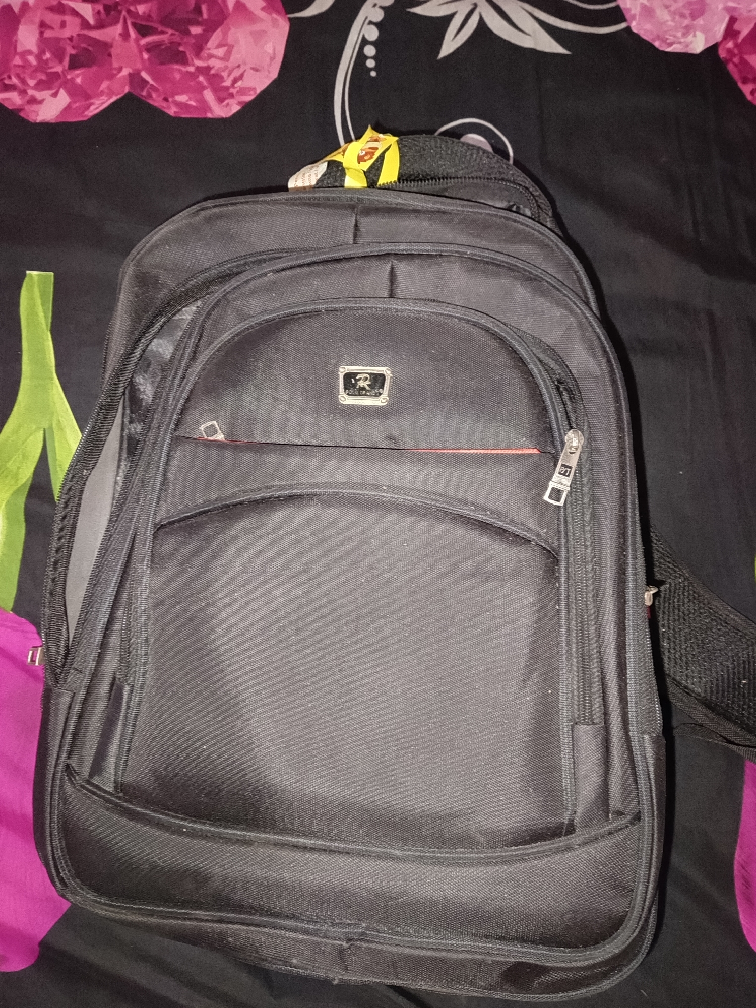 A black backpack is displayed on a black surface with floral patterns in pink and white. The backpack is the primary subject, oriented vertically and occupying the majority of the frame. It has multiple zippered compartments. A small yellow and red packaged item is partially visible at the top of the backpack, near the strap. The background is a patterned fabric, featuring large pink floral shapes at the top and green leaf-like shapes on the left side. The lighting is somewhat dim, casting subtle shadows. The image appears to be a close-up shot, focusing on the backpack. The specific location in Terbanggi Marga, Indonesia, is not depicted or inferable from the visual elements presented in the image.