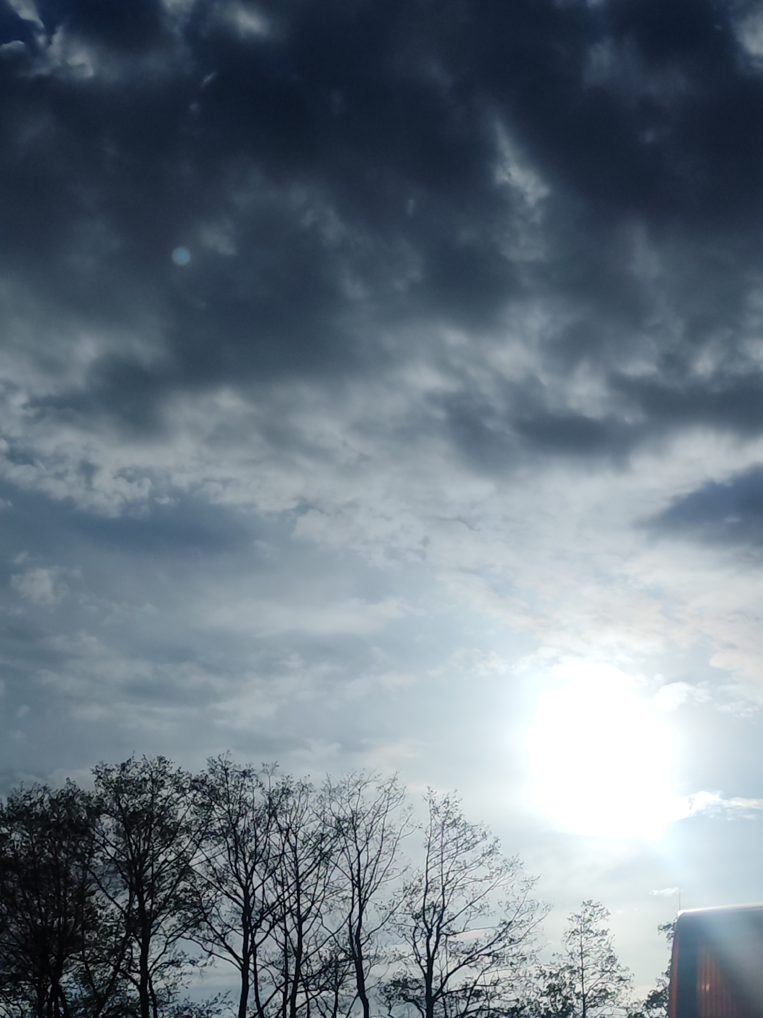 The image depicts a sky with dark, heavy clouds and a bright sun obscured by haze or thin clouds. In the lower part of the image, silhouetted bare trees are visible against the bright sky. To the right, a portion of a building or structure is present. The precise location in the city of Chaikovsky, Russia, is not discernable from the image alone.