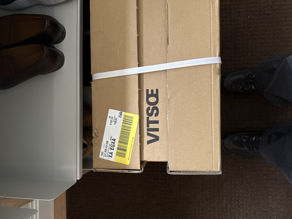 An overhead, slightly angled photograph captures an indoor scene, likely within a private residence or office in New York City, United States of America. In the foreground, the lower portion of a person's legs and feet are visible, wearing dark trousers and black lace-up shoes, positioned on a brown carpet. Directly in front of the person, two cardboard boxes are stacked horizontally. The top box, light brown in color, features a yellow and white FedEx shipping label adhered to its upper left corner. The label displays a barcode and tracking number "XA EGXA," along with "IP ECO CLR," "ETD CPL," "112.31," and the airport code "EWR," indicating routing via Newark Liberty International Airport, which serves the New York metropolitan area. A white fabric strap wraps vertically around both stacked boxes, securing them together. The lower box is also light brown cardboard and clearly displays the black block letters "VITSŒ" as a brand logo. Above the packages, a white, open-front shelf or storage unit is visible, beneath which a pair of dark brown loafers and part of a white sneaker are present on an upper surface, suggesting storage or shelving above the packages.