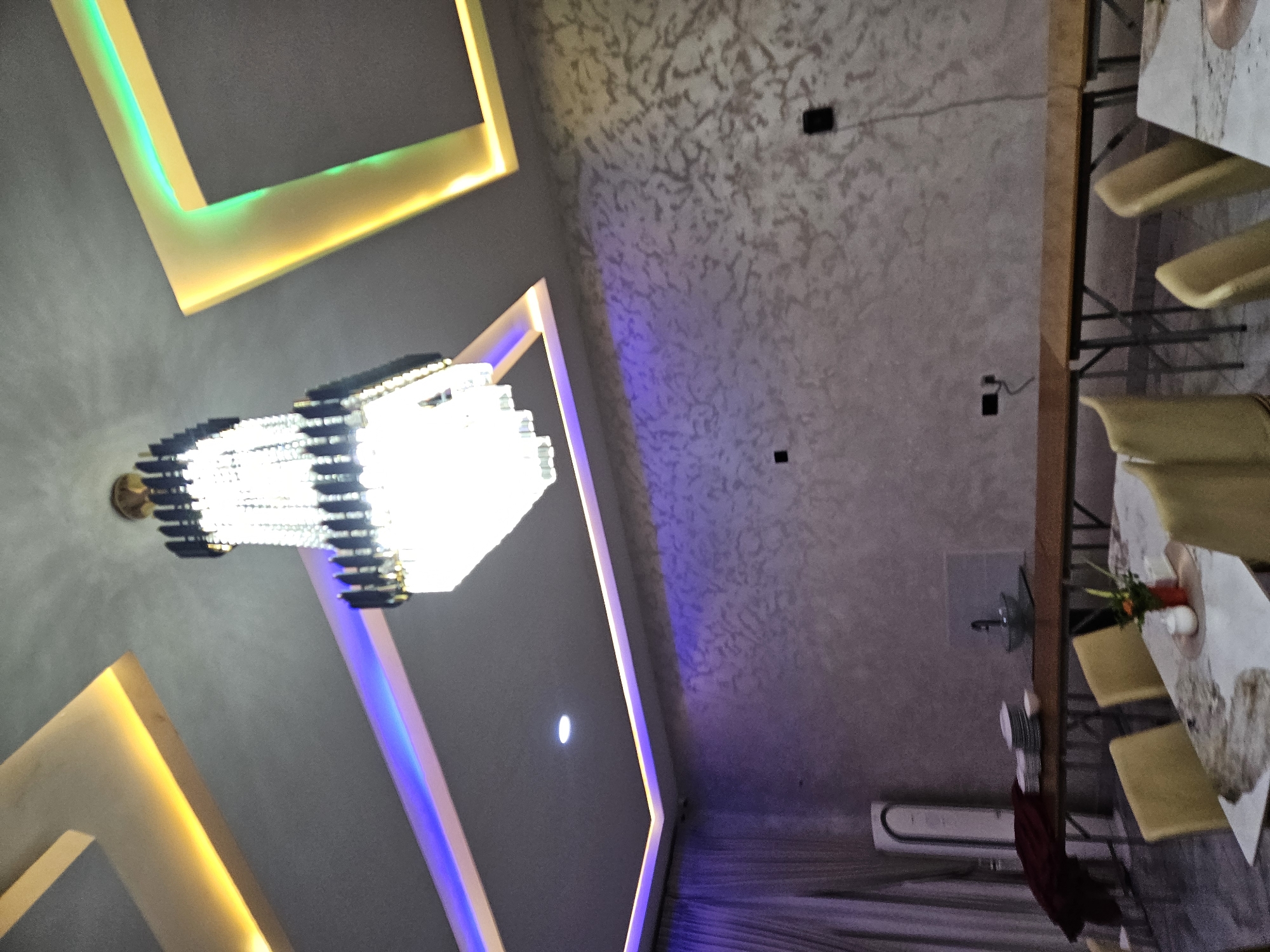 The image depicts an interior space in Akwanga, Nigeria, featuring a modern chandelier as a focal point, illuminated by bright white light. The ceiling design incorporates geometric shapes outlined with LED lighting, transitioning from green and yellow hues on the left side to blue on the lower portion. The ceiling has a textured finish in a neutral color. To the right, a table is partially visible, set with plates, silverware, and floral centerpieces, with chairs positioned around it. The room is illuminated by the chandelier, and there are visible electrical fixtures on the ceiling. Curtains are visible at the bottom of the frame.