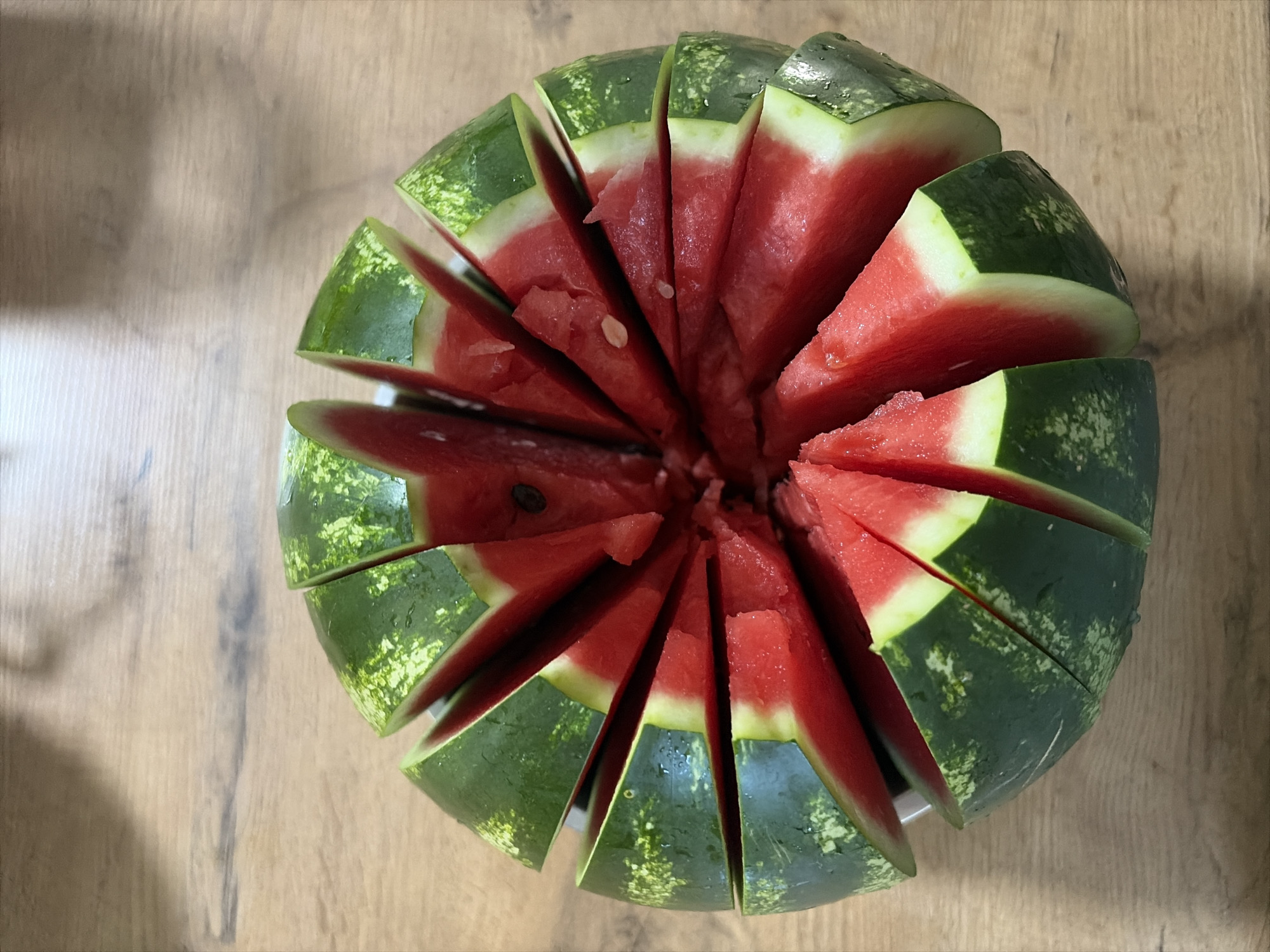 A whole watermelon, sliced radially into wedges, rests on a wooden table. The watermelon's green rind transitions to a bright red, juicy interior, with visible seeds in some slices. The scene is brightly lit, and the overall impression is one of freshness and a prepared treat. The location of Oxentea, Moldova, is not discernible from the image.