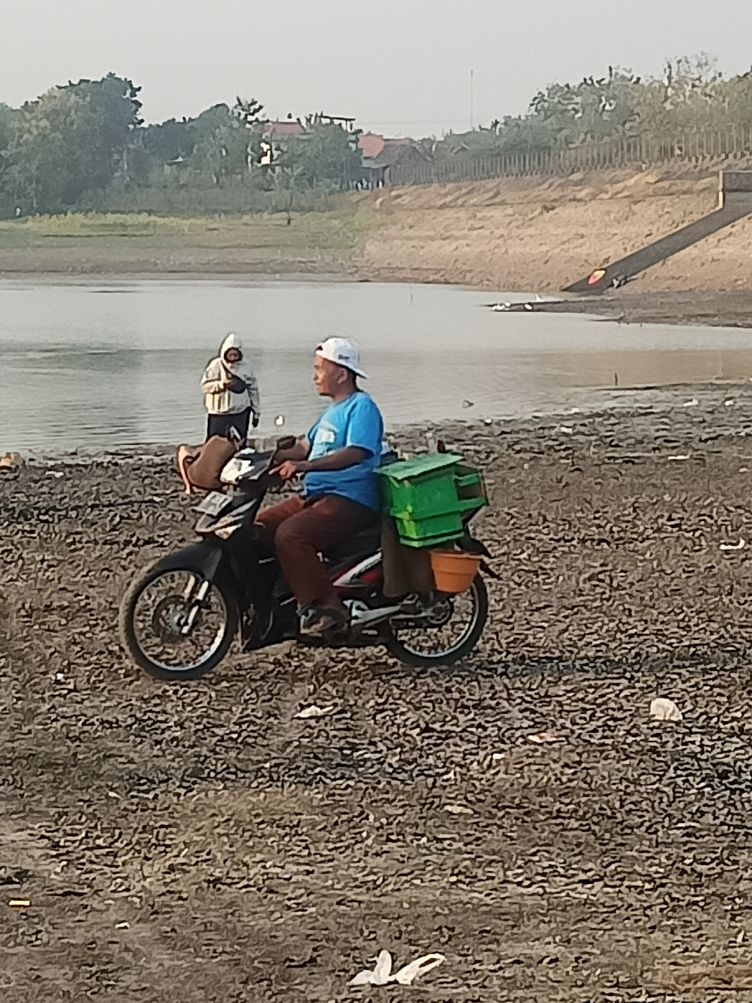 In Sambiduwur, Indonesia, a man wearing a blue t-shirt, brown pants, and a white baseball cap rides a black motorcycle. He is parked on a dry, rocky shore next to a body of water. Strapped to the back of his motorcycle are a green storage container and an orange bucket. In the background, another person wearing a white hooded jacket stands near the water's edge. The landscape includes trees and some buildings on a distant hill. The sun appears to be low in the sky, casting a warm light.