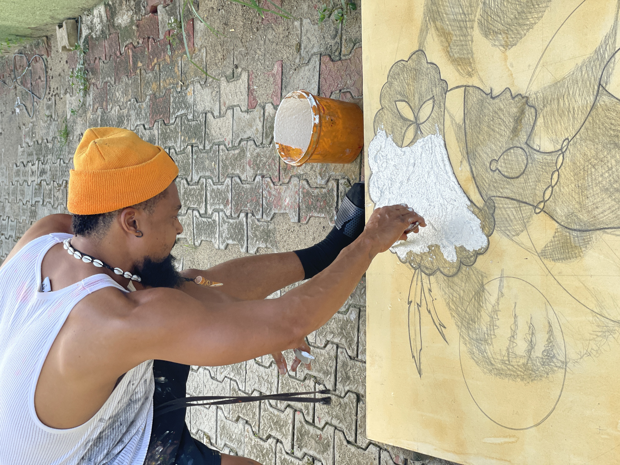 A man with a dark beard wearing an orange beanie, a white tank top, and a shell necklace stands in front of a cement wall. He is applying a white substance from an orange container to a large illustration of a person wearing a necklace on a yellow background. There are paintbrushes resting on the ground beside him. The location appears to be Ado, Nigeria, based on the streetscape.