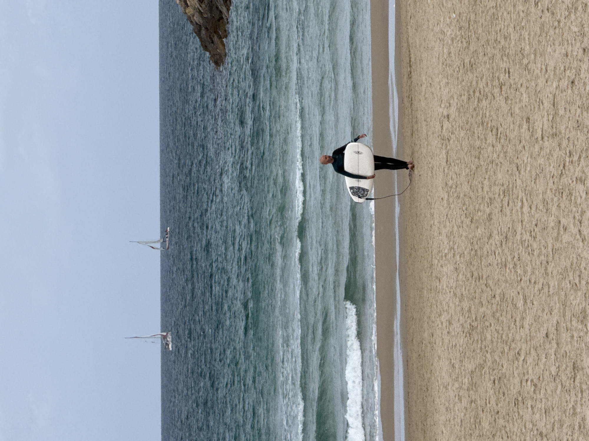 A surfer carrying a white surfboard walks on a sandy beach next to the ocean in Herzliya, Israel. Two sailboats are visible in the distance. A rocky headland is in the upper left corner of the frame.