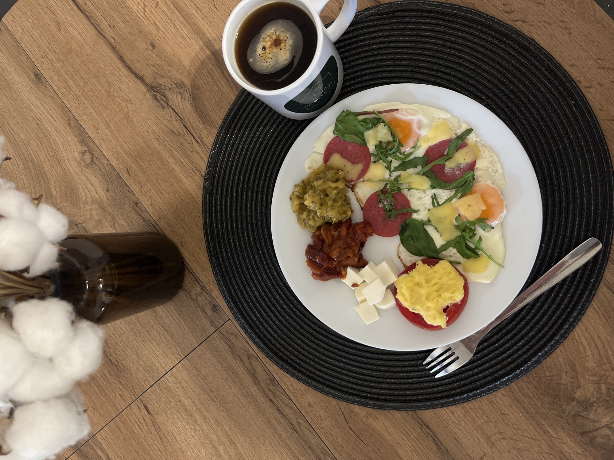A breakfast scene is captured in an overhead shot. A white plate sits atop a black circular placemat on a wooden surface. The plate holds eggs with sausage, cheese cubes, tomato topped with yellow food, green relish and stewed beans. A cup of coffee is next to the plate. A silver fork is to the lower right of the plate. A glass bottle with cotton sprigs is at the lower left. It is not possible to confirm the location of Литвиново, Russia.