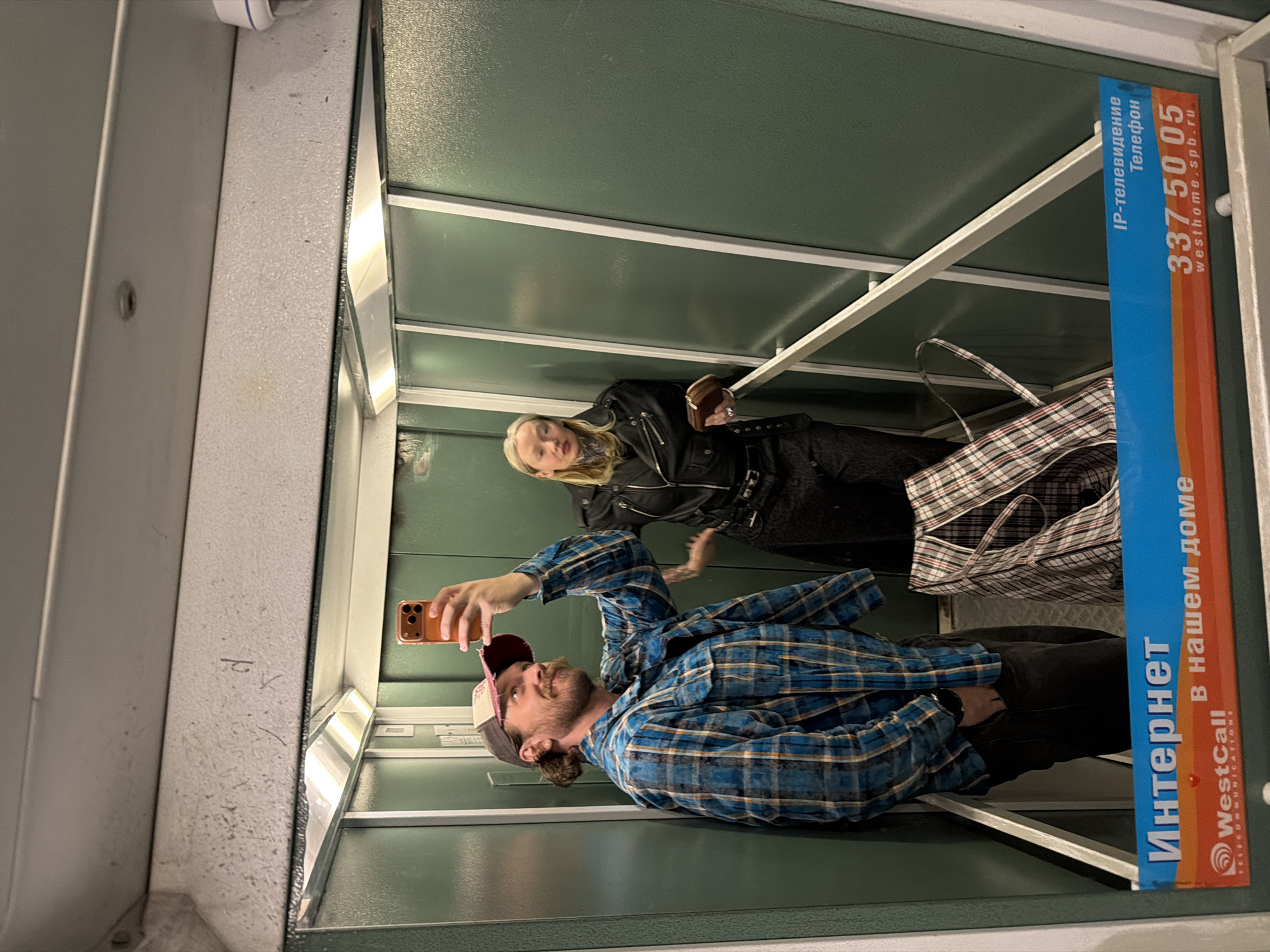 A man wearing a blue plaid shirt and a baseball cap is taking a selfie in an elevator. A woman in a black leather jacket stands beside him. An advertisement for "WestCall" Internet and TV services is visible on the elevator wall, with the phone number 337-50-05 and the website "westhome.spb.ru". The website address indicates that the location is likely in Saint Petersburg, Russia.