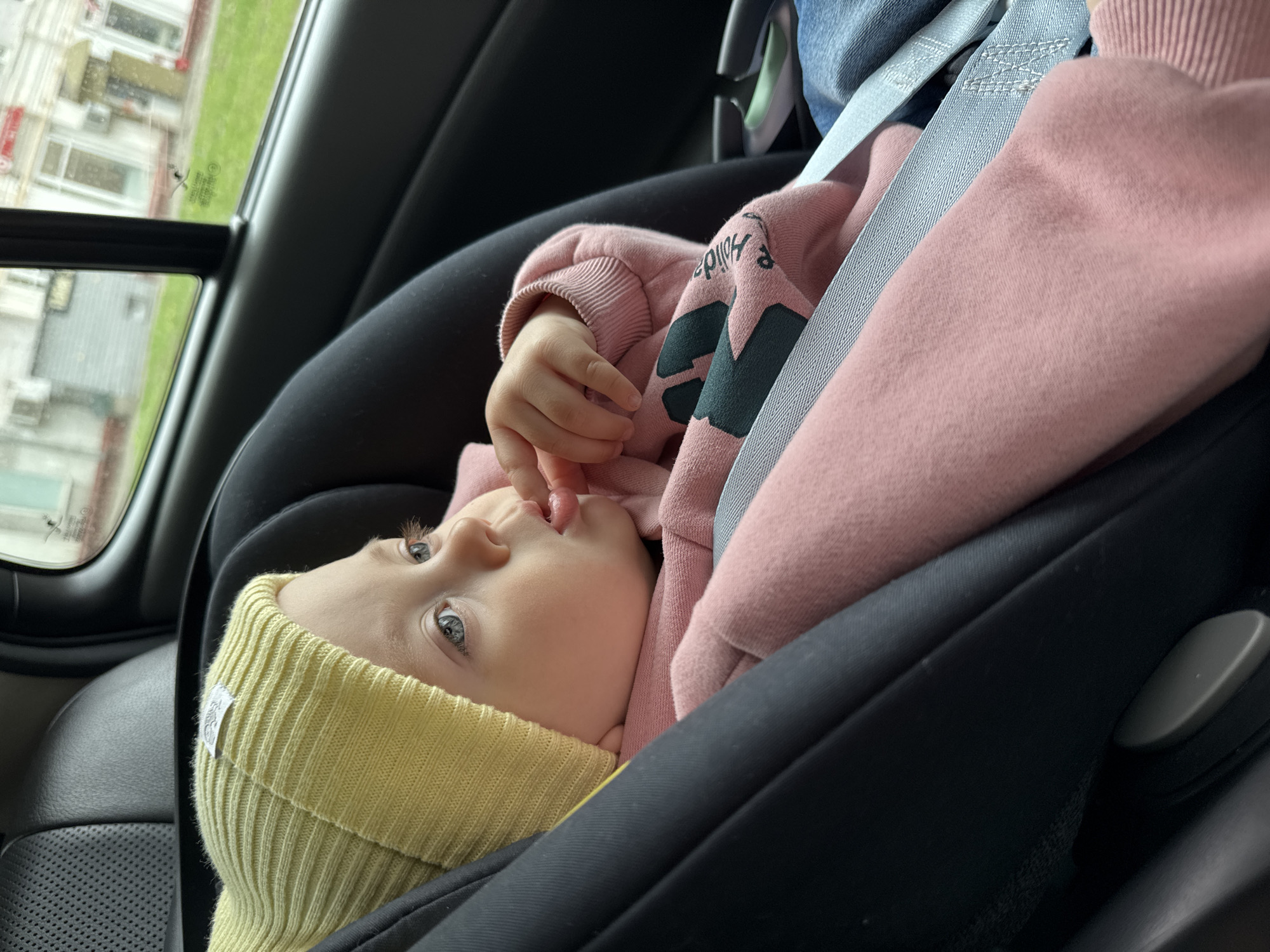 A baby in a car seat is shown in a close-up, angled shot. The baby is wearing a yellow knit hat and a pink sweatshirt with a dark green design. The baby is secured in a black car seat with grey straps. One of the baby's hands is near their mouth. A window is visible in the background, showing a building exterior. The location in the city of Moscow, Russia cannot be determined from this image.