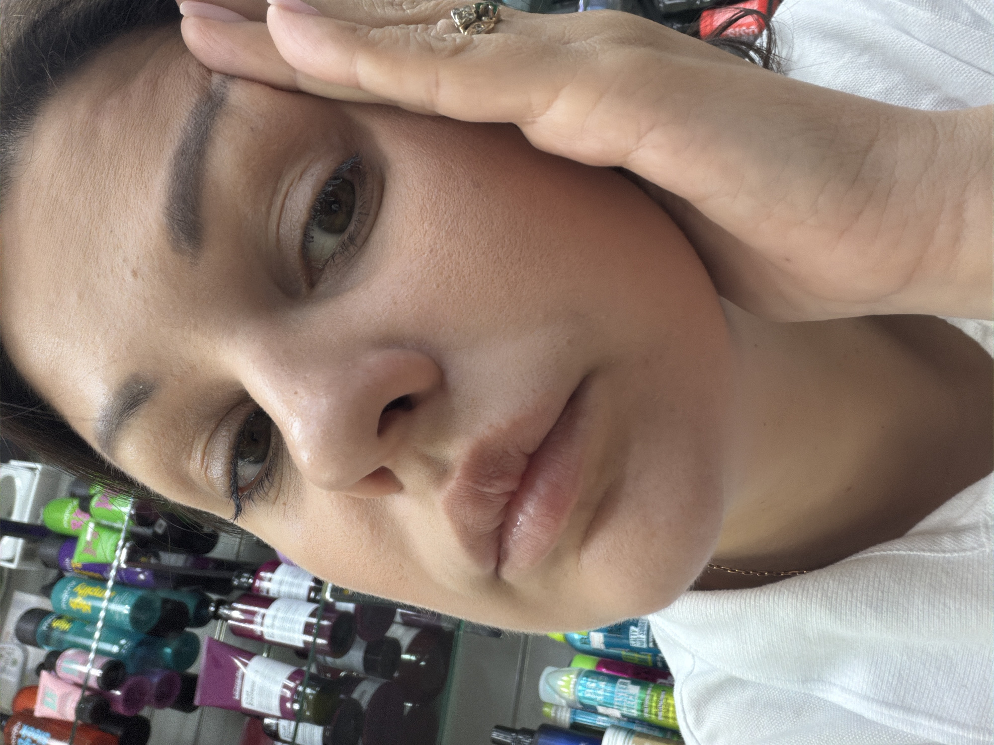 The image is a close-up, selfie-style photograph of a woman's face, taken indoors, likely in a retail environment such as a beauty supply store given the shelves of products visible in the background. The woman is looking towards the camera with her hand resting on her cheek. She has dark hair and is wearing a white collared shirt. She is also wearing a ring on her right hand. The specific location in the city of Sochi, Russia, cannot be determined from the image provided.