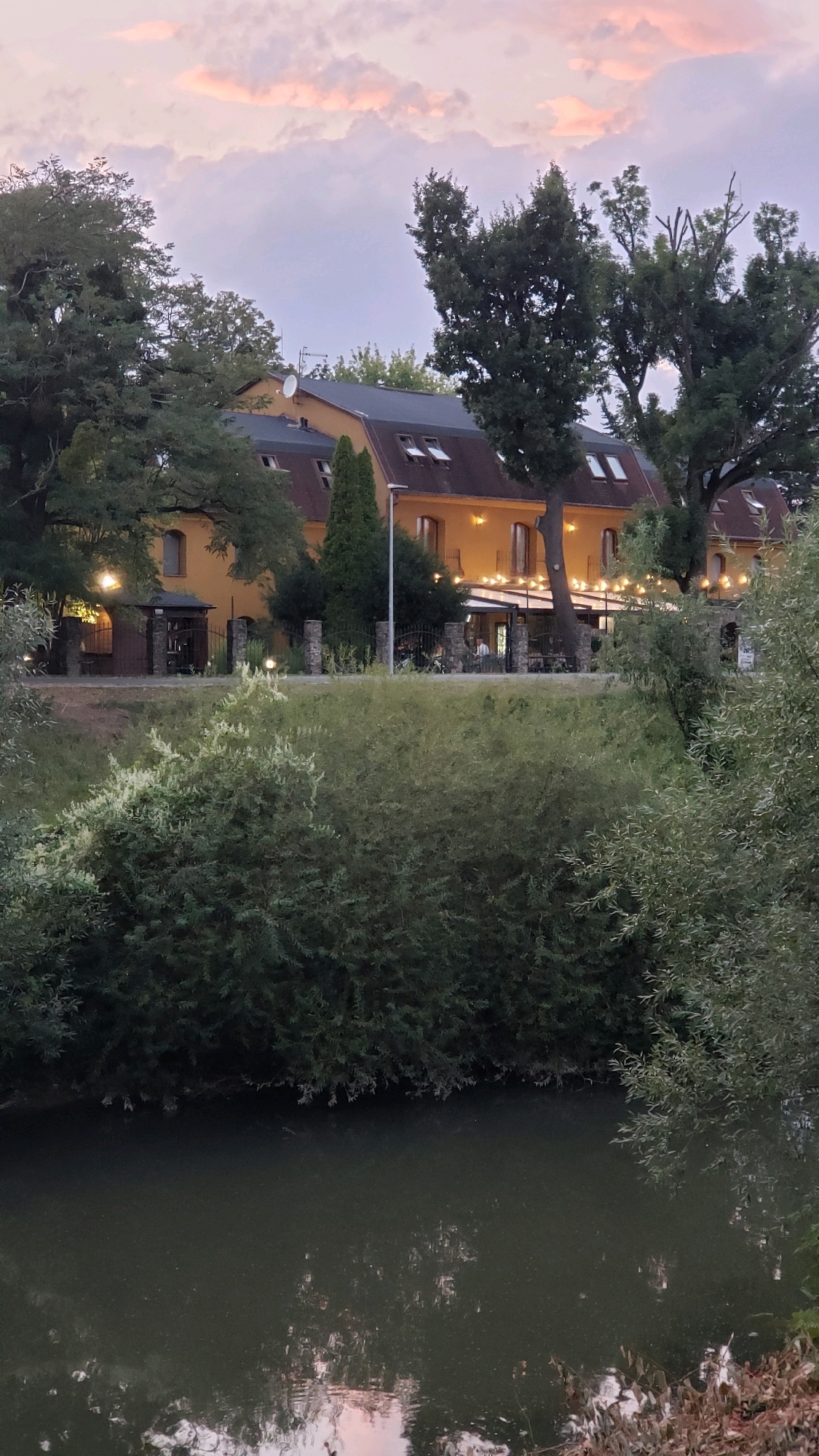 The image depicts a waterside building complex with an adjoining outdoor seating area, likely a restaurant or hotel, situated in Prešov, Slovakia. The building itself is a multi-story structure with a yellow facade and a dark roof. Numerous windows are visible, some with open shutters, and decorative string lights are illuminated, suggesting it is evening or dusk. Several trees are scattered throughout the property and surrounding landscape. In the foreground, a body of water with reflections of the sky and trees flows. A dense line of greenery separates the water from the building complex. The sky displays a gradient of colors, with pink and orange hues indicating sunset. Several people are seated at tables outside the building, interacting and dining.