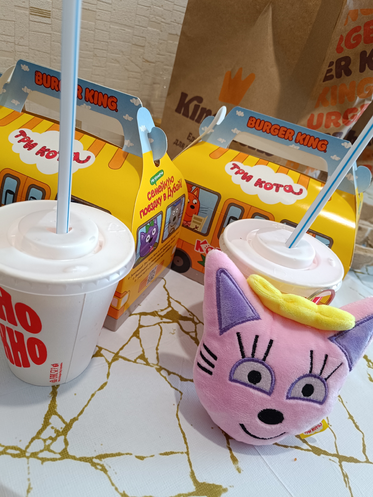 The image shows two Burger King kids meal boxes with "Три Кота" (Three Cats) branding in Russian, indicating they were purchased in Russia. Both drinks associated with the meals have straws protruding from their lids. A pink stuffed cat toy also rests on the table.