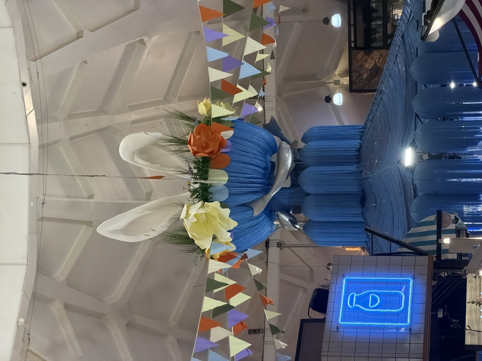 The image shows an interior space in Moscow, Russia, featuring an elaborate overhead decoration. The decoration includes blue and white floral arrangements and what appear to be stylized bunny ears, positioned among colorful triangular pennants. The surrounding environment consists of white, vaulted ceilings and structural supports, suggesting the interior of a large building or market. A bright neon sign with a bottle graphic is prominent on the right side of the frame. Additional architectural details, including light fixtures and what might be a section of roof or awning, are visible in the background. The image does not depict any people.