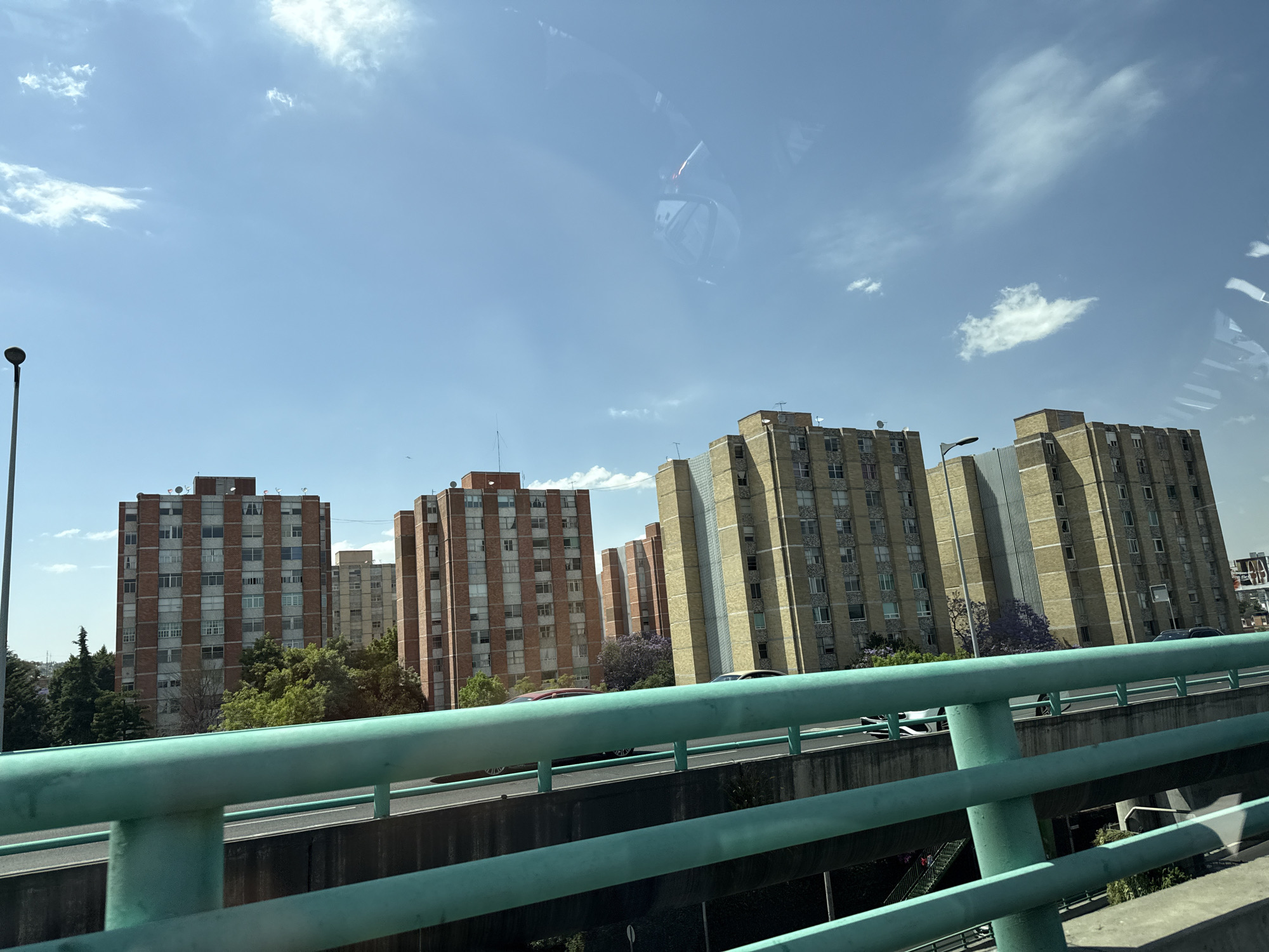 The image depicts a view from a vehicle driving along a highway in Mexico City, Mexico. The foreground is dominated by a light teal-colored railing, with the highway running below it. Beyond the highway, a series of tall, multi-story apartment buildings are visible. Some buildings have a pattern of red brick alternating with gray, while others appear to be primarily light yellow/tan with vertical gray stripes. There are trees and some purple flowering vegetation interspersed among the buildings. The sky is blue with scattered white clouds. The image is somewhat hazy, possibly due to the camera being behind glass. There are cars visible on the highway.