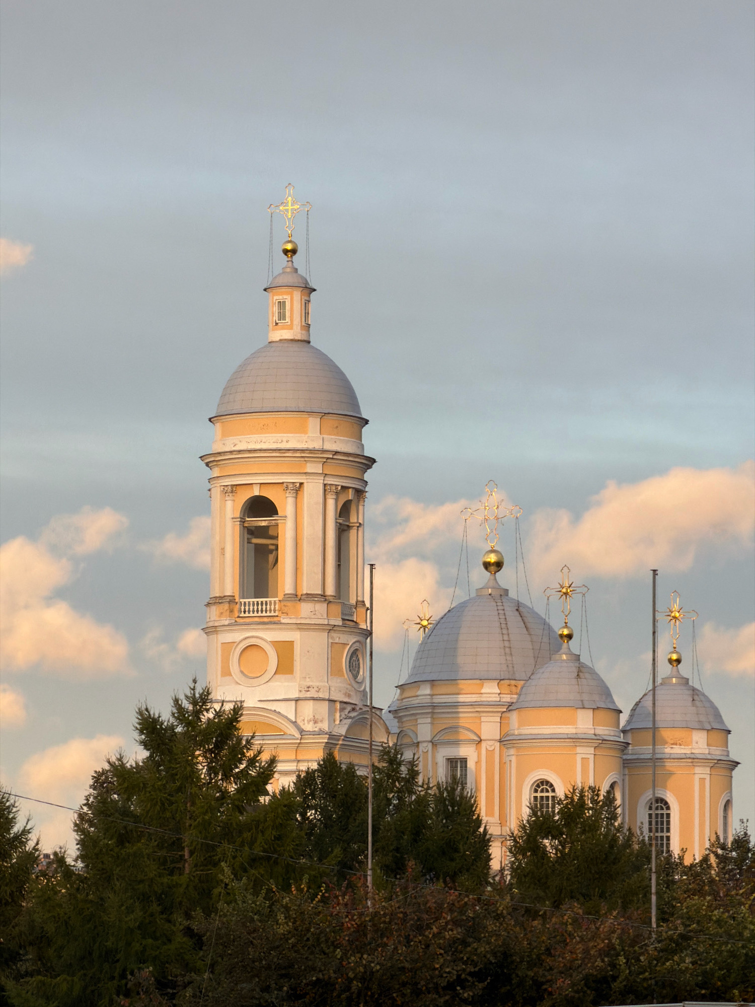 The image captures the bell tower and domes of the Alexander Nevsky Lavra in Saint Petersburg, Russia, against a backdrop of a cloudy sky. The architectural style features yellow facades, white decorative elements, and grey domes topped with ornate golden crosses. The bell tower, the tallest structure, has an open belfry with columns and a balcony. To its right, several smaller domes are visible, also adorned with crosses. In the foreground, dark green coniferous trees and deciduous trees with hints of autumn colors partially obscure the lower portion of the religious complex. The lighting suggests it is either early morning or late afternoon, with warm tones illuminating the clouds. There are no people or any discernible activity in the scene. The overall impression is of a serene, majestic religious landmark.