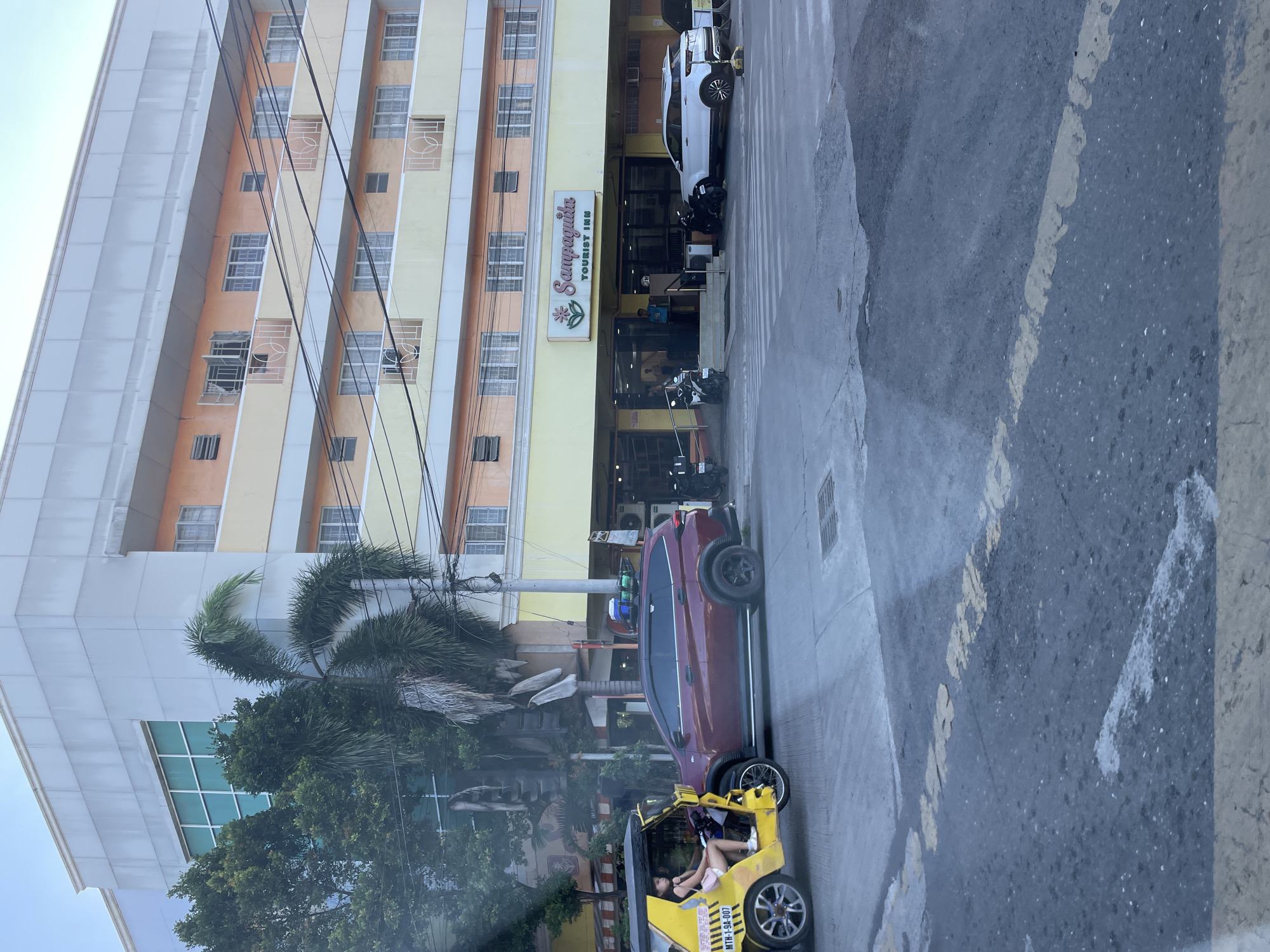 The image captures a street scene in Davao City, Philippines, focusing on a multi-story building housing the "Sampaguita TOURIST INN." The building's facade is characterized by a light grey upper section, with the lower floors featuring alternating bands of light peach and pale yellow, each containing multiple windows and small balconies with white, decorative metal railings.
At street level, the main entrance to the Sampaguita Tourist Inn is visible, identified by a prominent sign displaying "Sampaguita TOURIST INN" with a pink and green flower logo. A male individual in a light blue shirt stands near the inn's entrance. Parked directly in front of the inn are a white SUV (resembling a Range Rover Evoque) and several motorcycles. A pedestrian crosswalk with faded white stripes is visible on the asphalt road in front of the inn.
To the left of the frame, closer to the viewer, a yellow motorized tricycle with a sidecar is stopped. The tricycle displays the license plate "MTH-1-9A-007." A female passenger, wearing a light-colored top and shorts, is seated within the sidecar, facing forward. Adjacent to the tricycle is a dark red SUV, equipped with a roof rack holding various containers, also stopped on the road.
The street is paved with asphalt, showing faded yellow centerline markings and some white dashed lines. Lush green trees and several tall palm trees line the left side of the street, partially obscuring a utility pole with numerous black overhead wires and a traffic light. The sky above the building is clear and light blue.