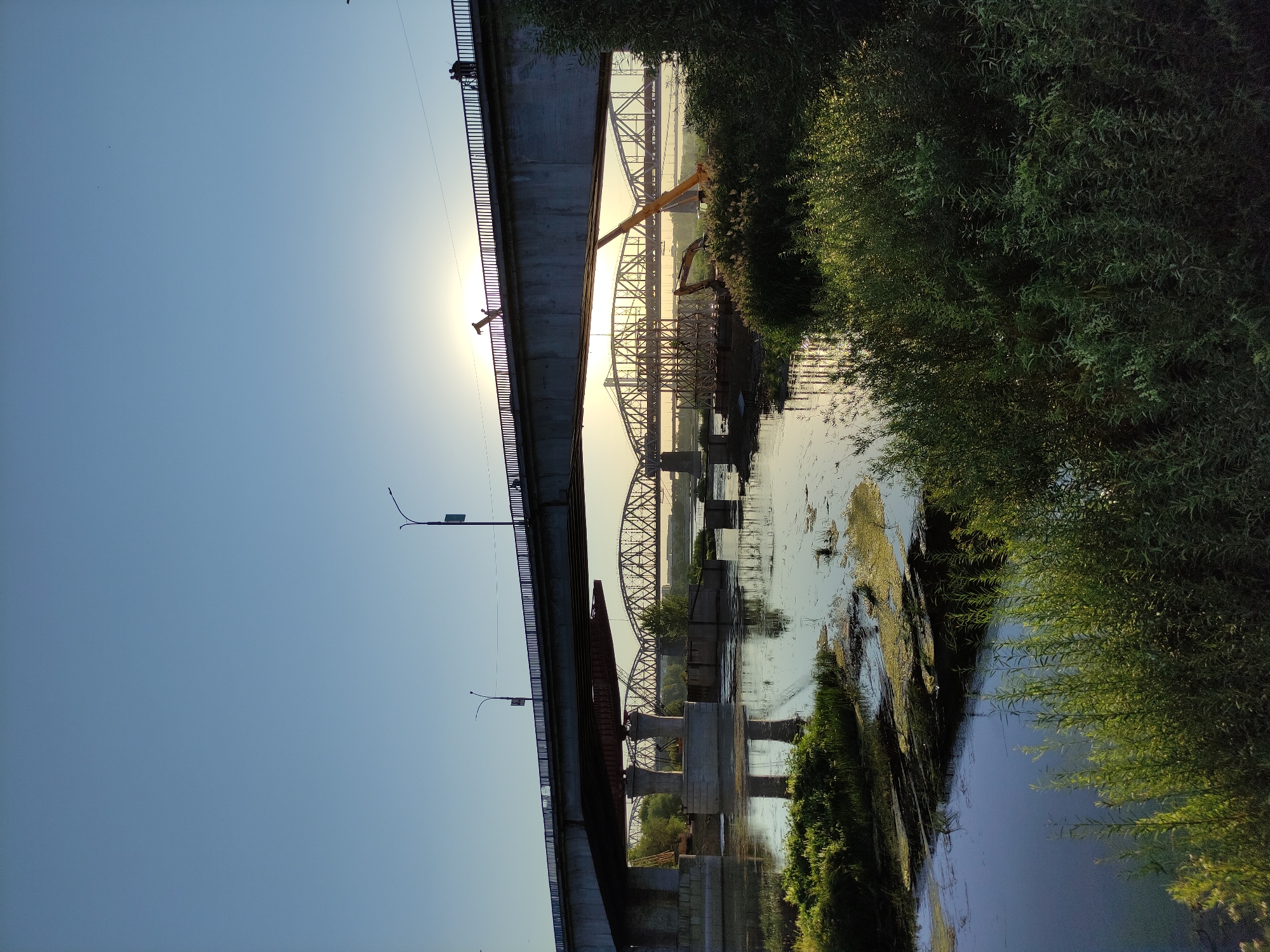 This image depicts a tranquil sunset scene over the Irtysh River in Semey, Kazakhstan. Multiple bridges span the river, with the foreground dominated by lush green vegetation and calm water reflecting the sky. In the mid-ground, construction is underway on one of the bridges, with cranes and building materials visible. Two silhouetted figures are walking on the top bridge, appearing to enjoy the sunset. The sky is clear with a gradient of blue, and the sun is low on the horizon, casting a warm glow over the scene.