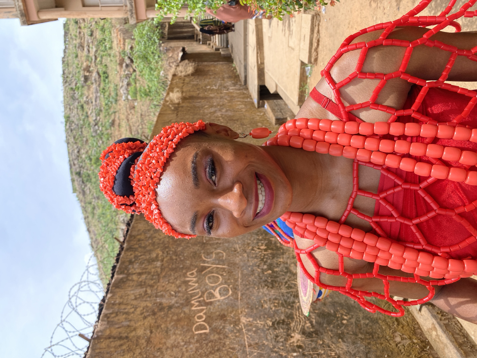 A medium, eye-level shot depicts a woman from the shoulders up, facing the camera and smiling. She is adorned in a traditional red beaded necklace and headpiece, likely of Igbo origin. She has dark hair, dark eyes, and makeup highlighting her eyes and lips.
She stands in front of a light brown concrete wall bearing graffiti reading "Damuwa Boys." Above the wall is a hill covered in vegetation, and to the right, a set of concrete steps and pathways lead uphill, with people visible in the distance. The location is likely in Gwong, Nigeria, given the clothing and scenery. Barbed wire is seen along the top of the wall, and the sky is visible above the wall and hill.