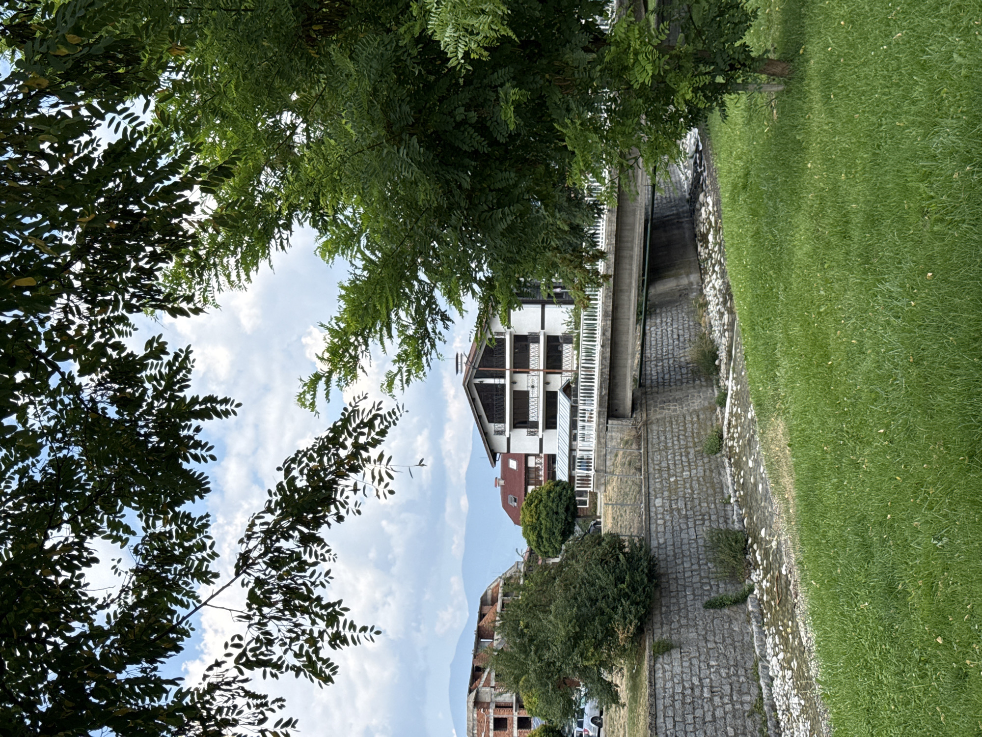 The image depicts a scene in Bansko, Bulgaria, featuring a small, shallow river with a rocky bed running through a grassy area. A concrete bridge spans the river, supported by a stone embankment. On the far side of the river, a white building with dark trim and multiple balconies is visible, along with other structures, some of which appear to be under construction. Lush green trees and foliage frame the scene, with some branches extending into the foreground from the top of the image, partially obscuring the sky. The sky itself is partly cloudy. The overall environment suggests a tranquil, possibly semi-urban setting with a natural element.