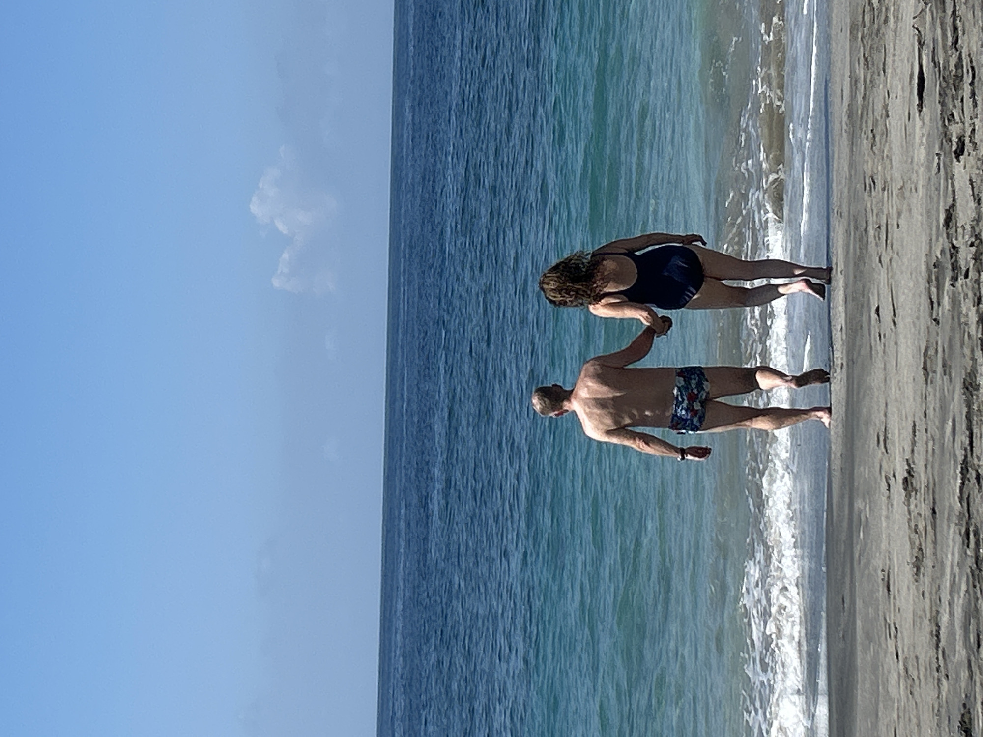 A couple, a man and a woman, walk hand-in-hand on a sandy beach towards the turquoise ocean under a bright blue sky with scattered clouds. The man, from behind, wears patterned swim trunks. The woman, also from behind, wears a dark blue one-piece swimsuit. Gentle waves lap at the shore. The scene depicts a serene coastal environment.