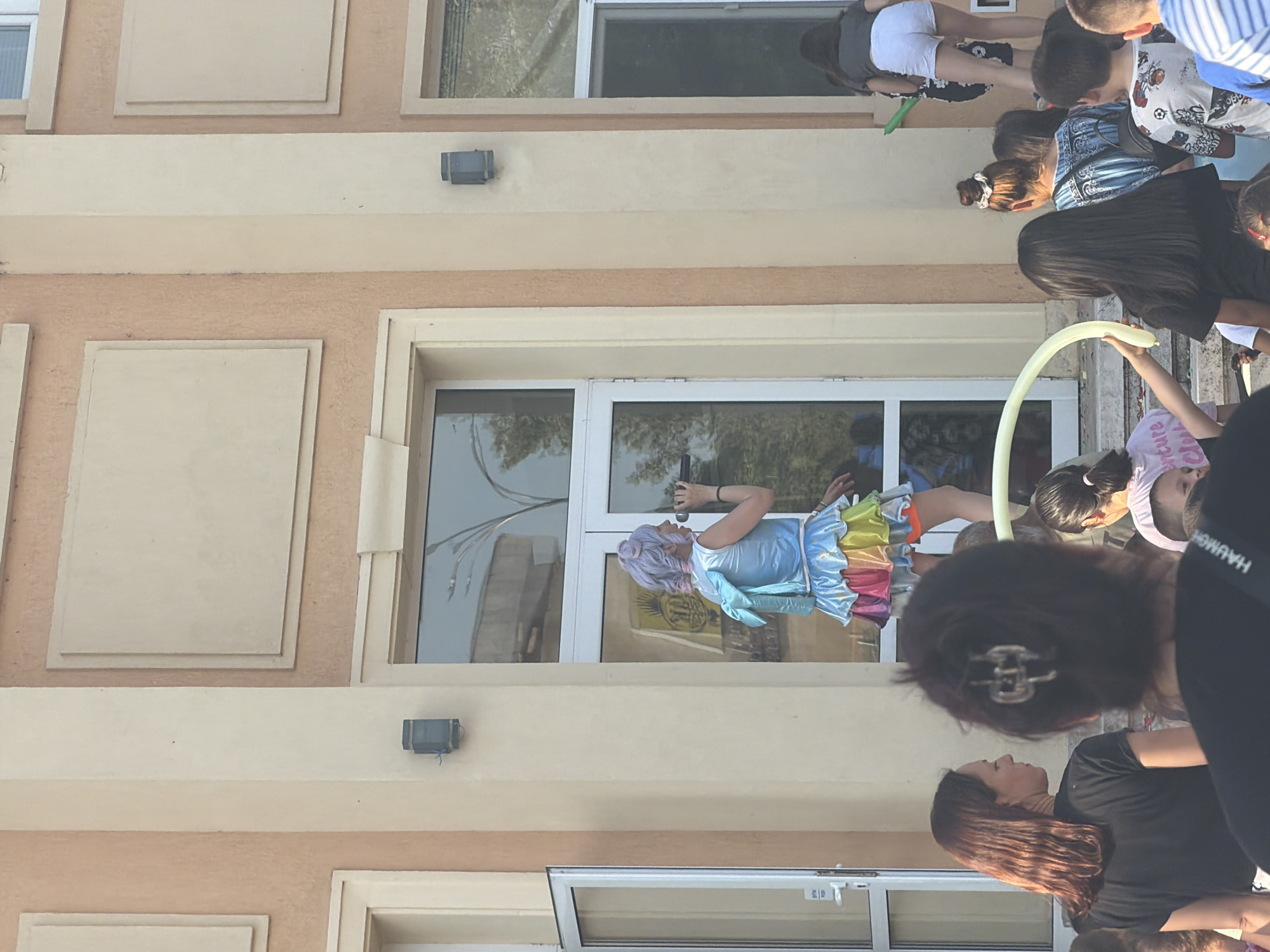 Here's a description of the scene:
A medium-angle shot shows a gathering of people outside a two-story building. The exterior of the building is primarily a light beige color, with some sections painted a slightly darker shade of beige. There are rectangular decorative features on the facade. The building has large windows, some of which reflect trees.
A woman in a fairy-like costume, with purple hair, a light blue top, and a colorful rainbow skirt, is standing near a window. She appears to be taking a photograph with her phone. A group of people stands around her, some looking at her and others interacting with each other. One person holds a long, yellow balloon. The foreground shows a woman with dark hair and a black shirt. The location is in an area of Unknown, Unknown.