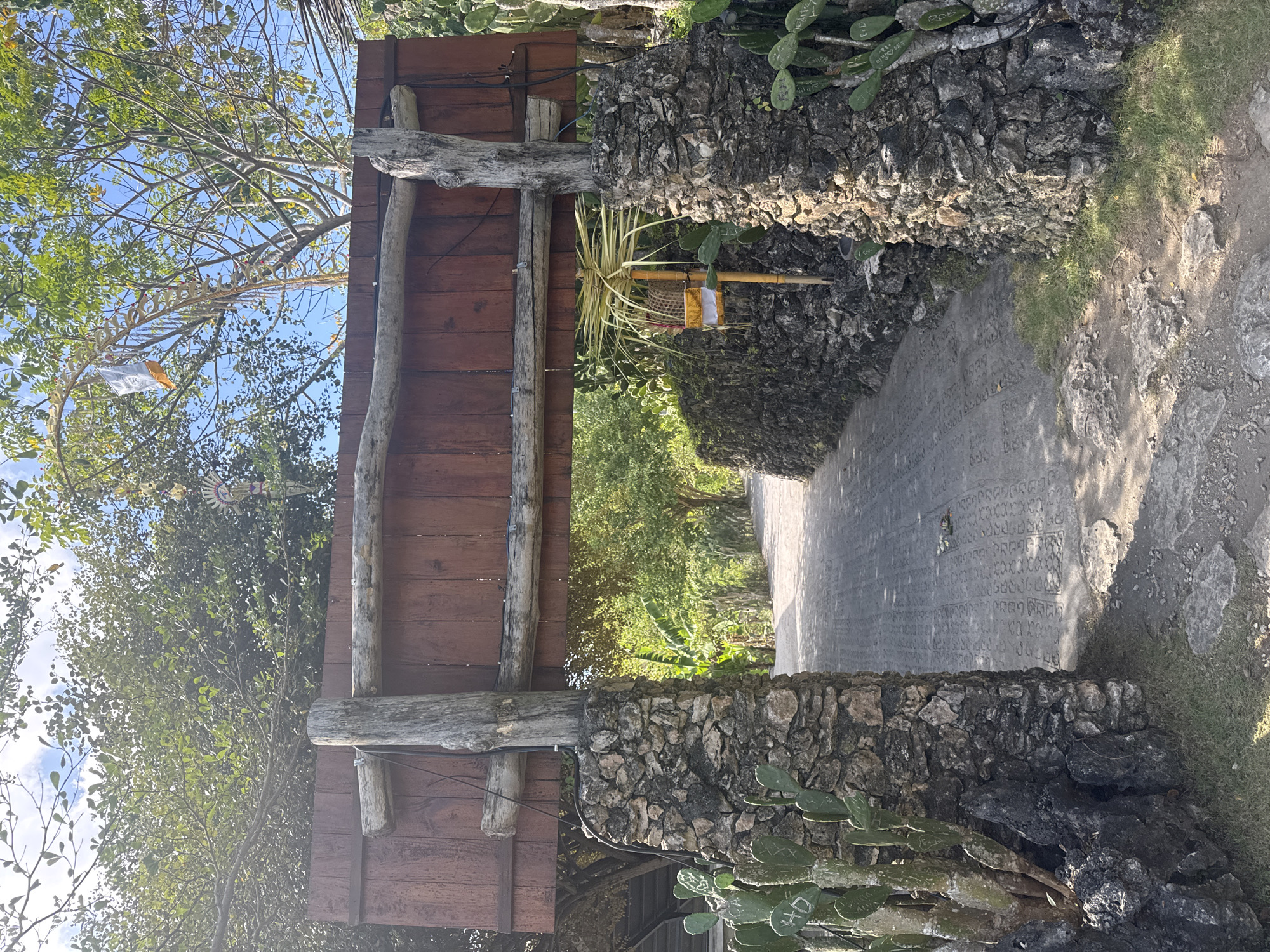 An outdoor pathway in Nusa Penida, Indonesia, is framed by an arched gateway constructed of stone pillars and a wooden plank sign supported by rough-hewn logs. The pathway, paved with patterned concrete tiles, leads into a lush green area with trees and foliage. Cactus plants grow alongside the stone pillars, and a small, traditional offering basket is visible on a post to the right. The sky above is blue with scattered clouds, and the scene is illuminated by sunlight.