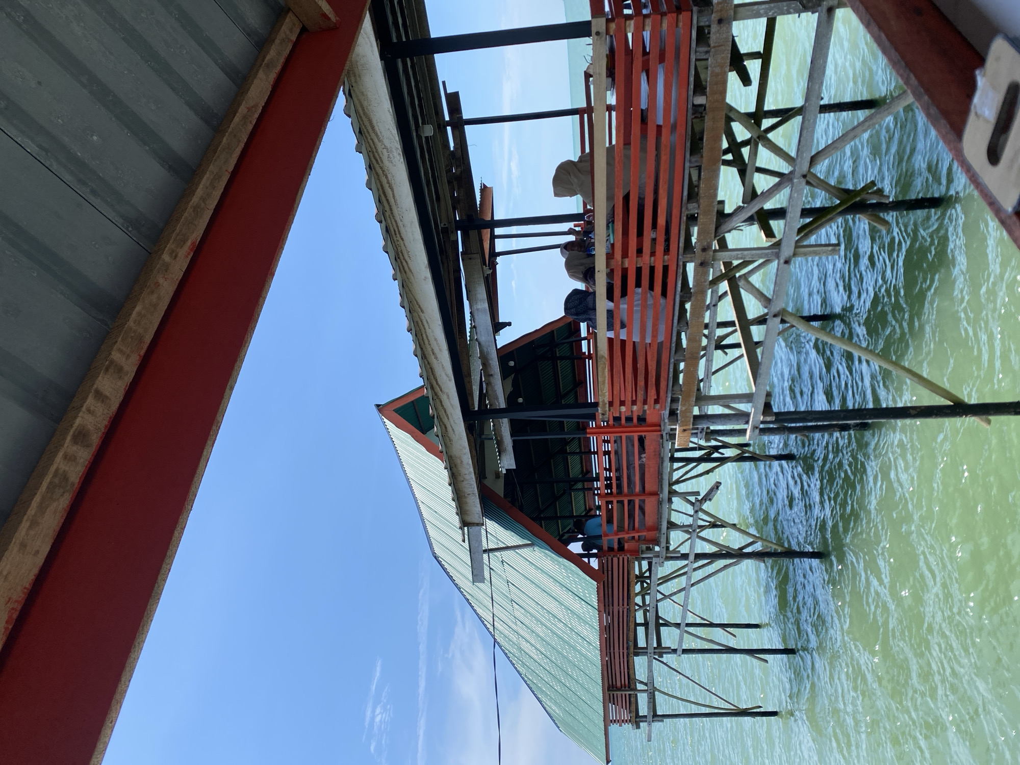 An over-the-water establishment is pictured in Lancok, Indonesia, on a clear, sunny day. The structure, built on stilts over the turquoise water, features a prominent green, corrugated metal roof with red trim. A wooden deck with red railings surrounds the open-air seating area where four individuals are seated at tables. The water beneath the structure is calm, with ripples indicating gentle movement. The sky is a vibrant blue with a few wispy clouds. The overall scene depicts a relaxed, waterfront dining or gathering place.