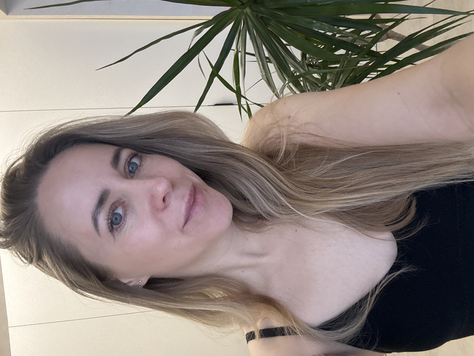 A selfie captures a woman with long, light brown and blonde hair, which cascades over her shoulders and chest. She has fair skin, prominent blue eyes, and a calm, slightly smiling expression as she looks directly at the camera. She is wearing a simple black top with thin straps.
The setting is indoors, featuring a light-colored wall or furniture in the background, suggesting a clean and minimalist environment, likely a home. A green houseplant with long, thin leaves (possibly a Dracaena) is visible behind her head and shoulders, adding a natural element to the scene. The lighting appears soft and natural, indicative of an indoor daytime shot. There is no visible text. The overall mood is relaxed and casual. This indoor scene is situated in Chaikovsky, Russia.