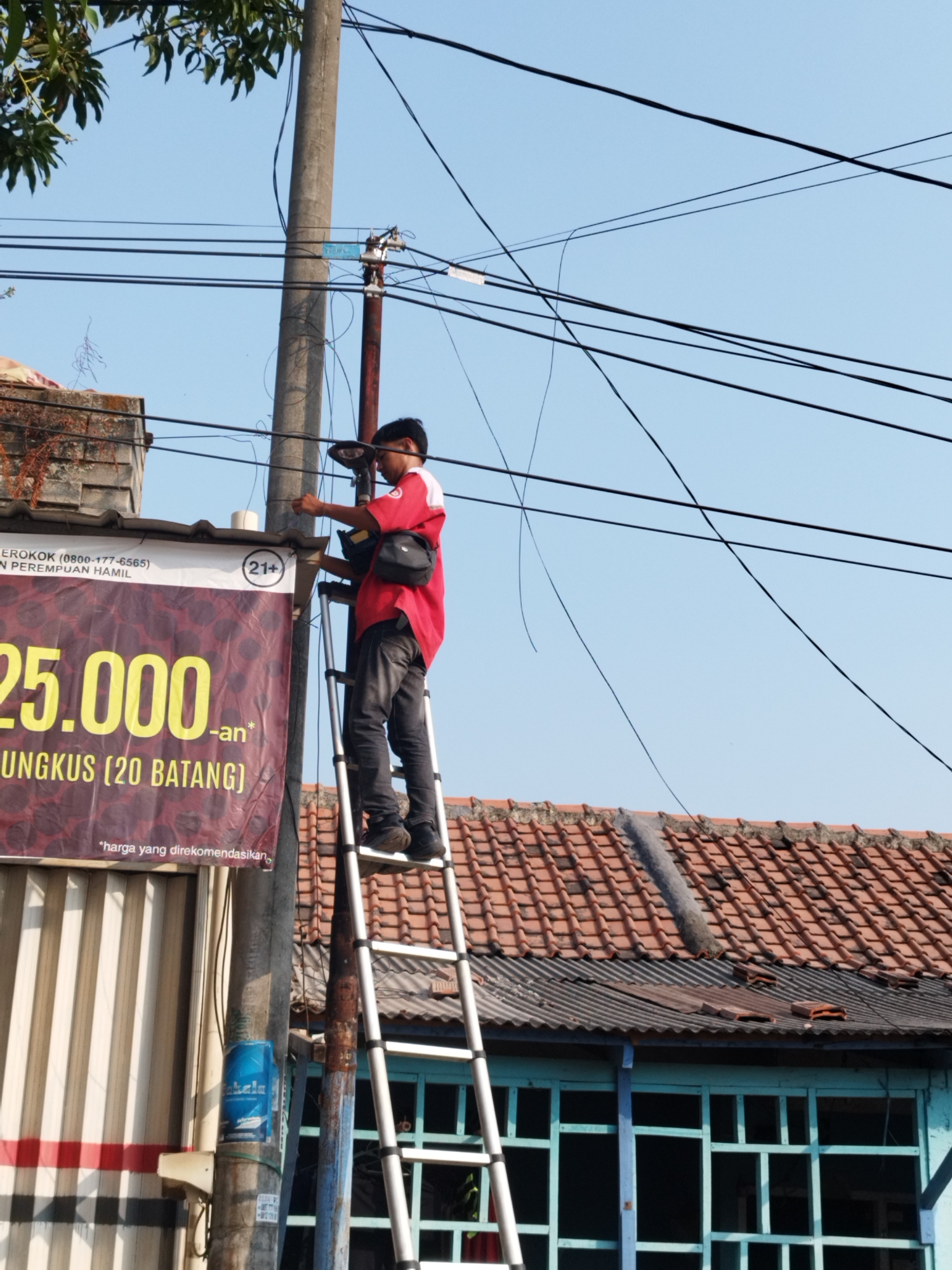 A man in a red shirt and dark pants is on a ladder, working on overhead wires next to a utility pole. He is holding tools and appears to be engaged in electrical work. A large banner with yellow text and a "21+" symbol is visible to the left of the utility pole. The background shows a tiled roof and a building with blue trim. The scene is outdoors with a clear blue sky. The banner mentions "KARAWANG" and "ROKOK (0800-177-6565) N PEREMPUAN HAMIL," which suggests this location is in Karawang, Indonesia, and the banner might be related to a local advertisement or public health warning concerning cigarettes and pregnant women.
