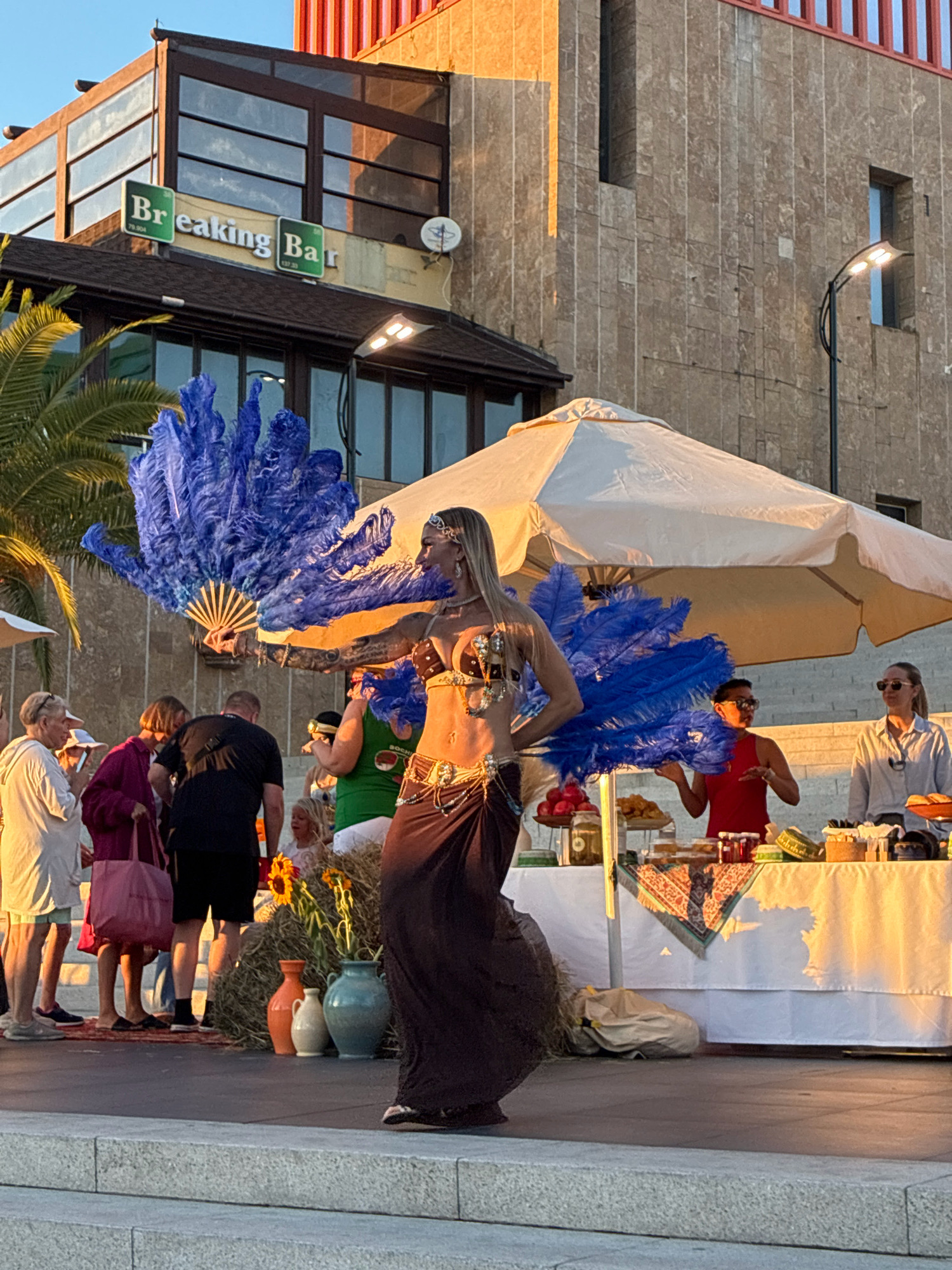 In Sochi, Russia, a woman dressed as a belly dancer performs outdoors. She has blonde hair and wears a gold sequined bra and a dark brown skirt. Large blue feather fans are held in her hands. Behind her, a white tent covers a table with various items for sale, including fruits, jars, and food. Several people are gathered, watching the performance and browsing the items. The background shows a building with a sign that reads "Breaking Bar" and a stone-tiled wall, illuminated by the warm glow of the setting sun. A palm tree is visible to the left of the scene.