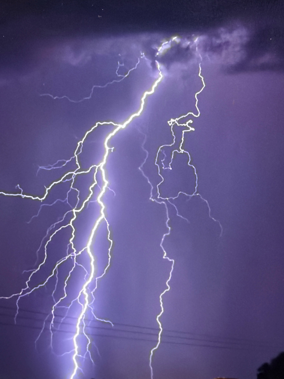 The image shows a dramatic lightning strike during a nighttime storm over the city of Kafr Yasif, Israel. A bright, jagged bolt of lightning illuminates the dark purple sky, branching out into multiple smaller streaks. Wispy clouds are visible, tinged with the purple hue of the storm. In the lower portion of the image, faint outlines of power lines and some dark foliage suggest the presence of the city's landscape beneath the tempest. No people or specific identifiable structures are clearly visible.