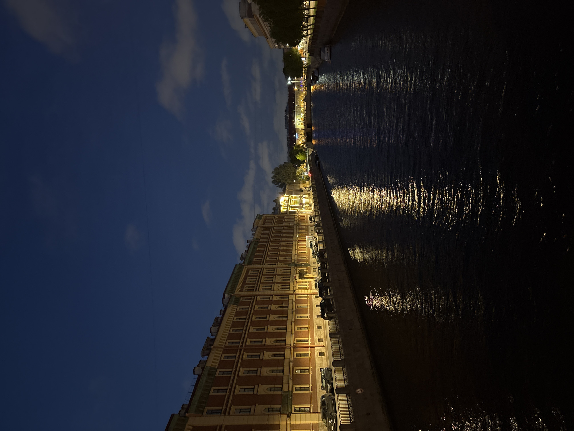 The image depicts a night scene along a canal in Saint Petersburg, Russia. On the left side, a large, ornate building with multiple floors and numerous windows is illuminated. The building is constructed of a reddish-brown material with detailed architectural features. Along the building's base, a sidewalk is visible, with parked cars lining the street. A railing separates the sidewalk from the canal.
The canal itself is dark, with reflections of the city lights shimmering on the water's surface. The right side of the image is predominantly dark, showing the water stretching into the distance. In the far distance, more buildings and lights can be seen along the opposite bank of the canal. The sky above is a dark blue, with scattered clouds illuminated by the city lights.