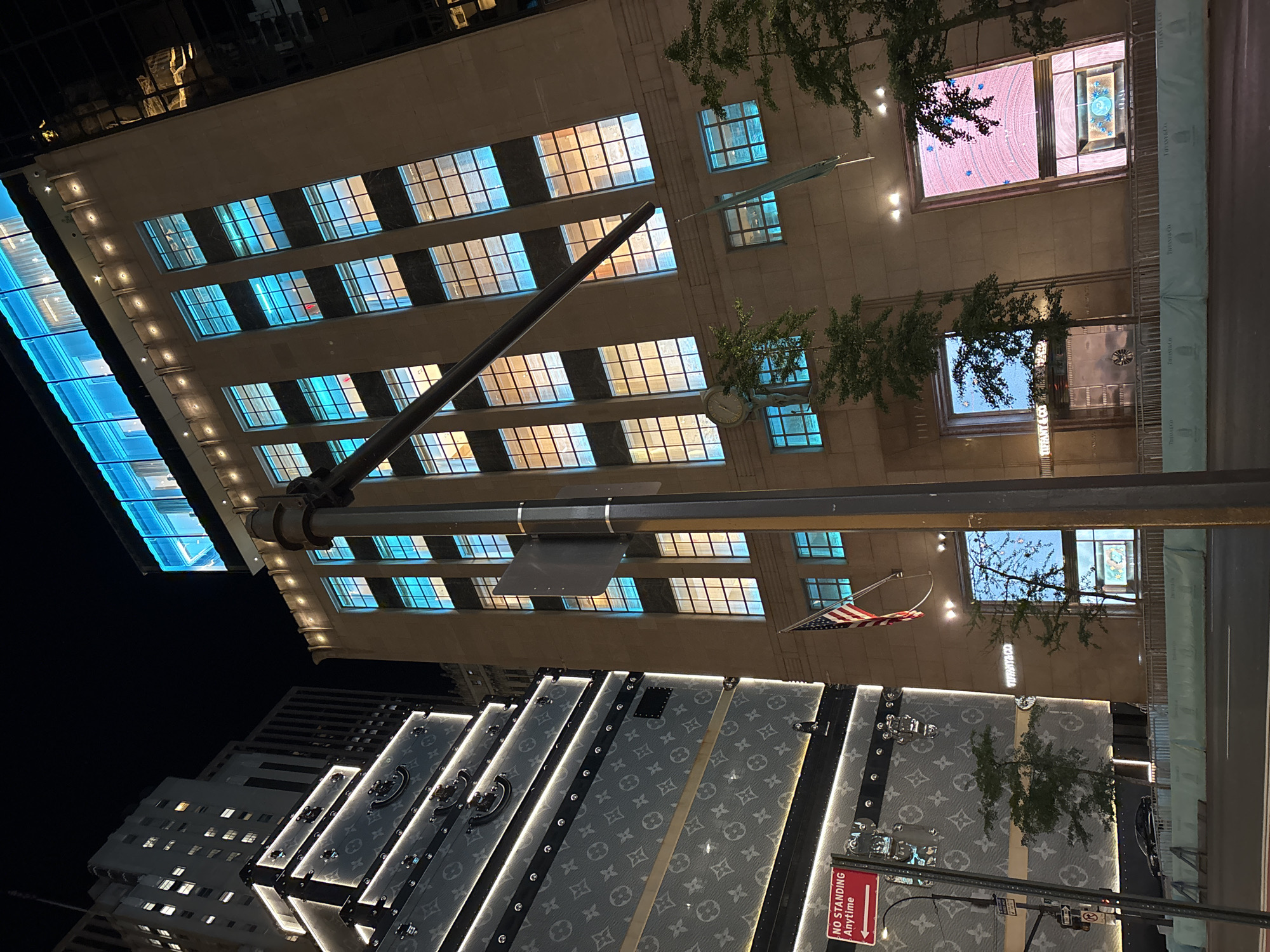 This nighttime image captures a vibrant urban street scene, focusing on the illuminated facades of prominent buildings. The perspective is tilted, looking upwards at the structures from street level.
The most prominent building, occupying much of the upper right portion of the frame (when viewed upright), is a large, light-colored edifice featuring numerous grid-patterned windows. Many of these windows are brightly lit, some with a warm yellow glow and others with a distinct cool blue light, creating a striking contrast. Green foliage from trees is visible in front of this building. Along its street level, light blue banners or fencing repeatedly display "TIFFANY&CO" branding, identifying it as a Tiffany & Co. store. An American flag is also seen hanging from this building.
In the lower left foreground, large, decorative structures resembling oversized Louis Vuitton trunks are brightly illuminated. These trunks showcase the iconic Louis Vuitton monogram pattern, adding to the high-end retail atmosphere. Behind these, another building with some lit windows can be seen against the dark night sky.
Visible text includes "TIFFANY&CO" on the light blue barriers and a red "NO STANDING Anytime" sign attached to a pole near the Louis Vuitton trunks. The dark sky confirms it is nighttime, and the artificial lighting casts a dramatic glow over the scene. No people are visible, allowing the focus to remain on the architectural details and luxury branding. The combination of Tiffany & Co. and Louis Vuitton suggests a prime commercial district, likely in New York City.