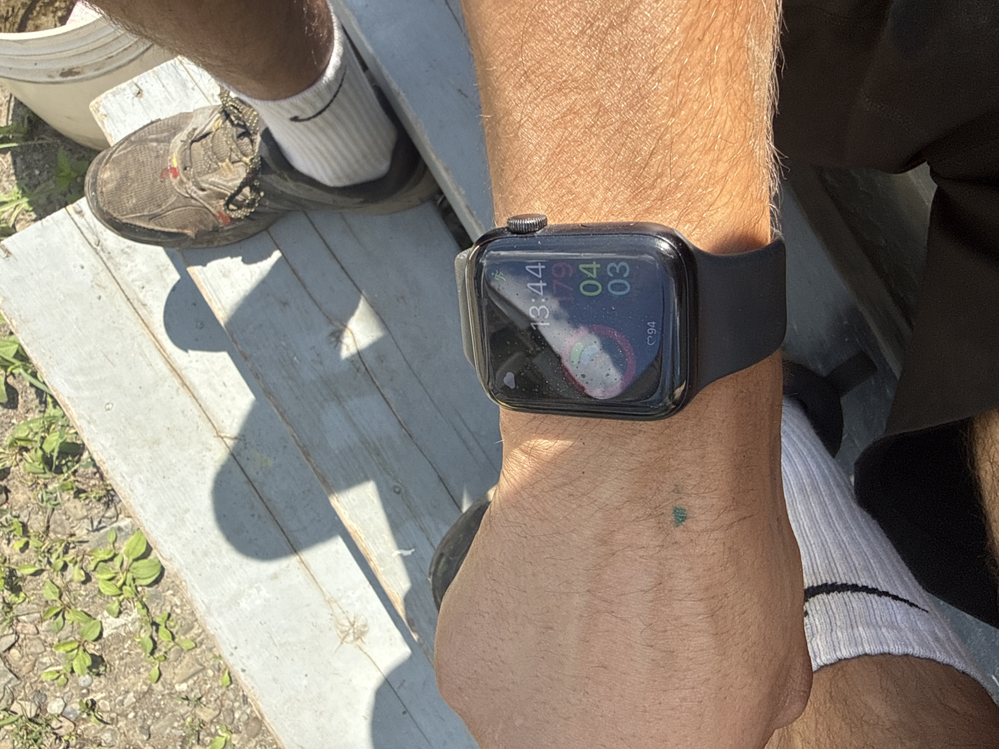 A close-up, outdoor, over-the-shoulder shot focuses on a person's left wrist, which is adorned with a black Apple Watch. The watch face displays the time as 13:44, along with activity rings for "move," "exercise," and "stand" goals, showing completion of 179, 04, and 03, respectively. The watch band is black and made of silicone. The person's skin is tanned and shows some body hair. A small, faded green tattoo of what appears to be a stylized tree or leaf is visible on the wrist. The background shows a weathered wooden surface with sunlight casting shadows. To the left of the wrist, part of a worn, tan shoe with brown laces and a white sock is visible. The lower left portion of the image reveals a patch of dry ground with scattered small green weeds. The scene appears to be in the Village of Johnson City, United States of America, likely in a backyard or outdoor recreational area due to the casual attire and natural elements.