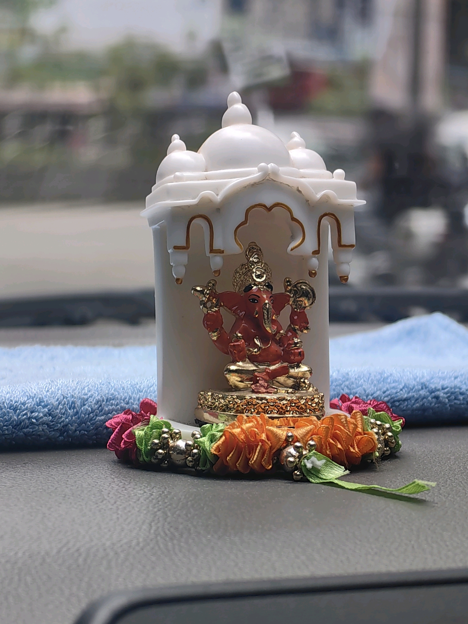 An indoor shot captures a small, decorative idol of Lord Ganesha placed inside a white miniature temple structure. The idol is red and gold, adorned with jewelry. The temple has a white dome-like roof with gold trim. A colorful floral garland, featuring pink, orange, and green ribbons with small silver beads, encircles the base of the temple. Behind the idol and temple, a soft blue cloth is visible. The background is blurred, suggesting an indoor setting, possibly a dashboard or shelf. The overall scene is static and does not depict any ongoing activity or discernible location within Mumbai.