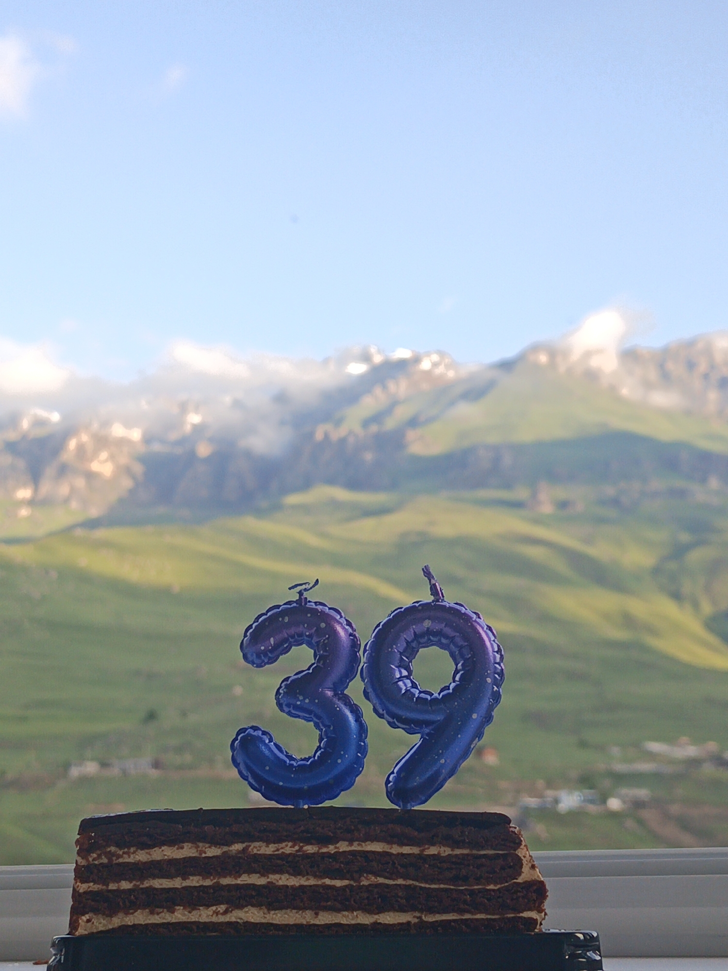 In Verkhny Fiagdon, Russia, a layered chocolate cake with visible cream filling sits on a dark cake stand. Perched atop the cake are two blue, foil balloon numbers: a "3" and a "9", signifying the number thirty-nine. The cake and balloons are positioned in the foreground, against a blurred backdrop of lush green mountainous terrain under a bright blue sky with scattered white clouds. The scale of the mountains suggests a natural, possibly remote setting within the region.