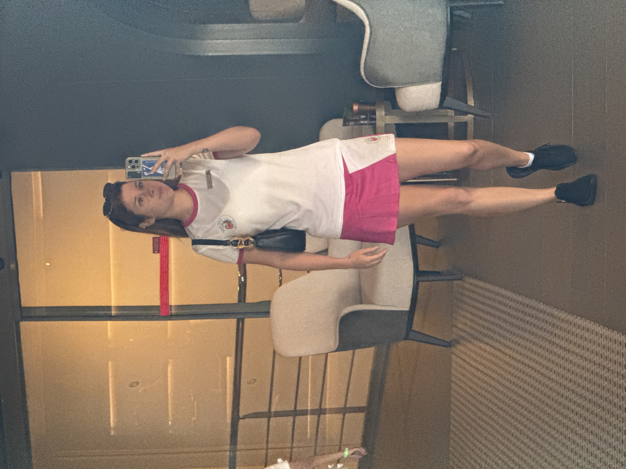A young woman with brown hair, wearing a white t-shirt with pink trim and a pink skirt, poses for a selfie in a hotel lobby. She is holding a phone in her right hand and has sunglasses pushed up on her head. She is wearing black sneakers and white socks. A black purse is slung over her left shoulder. The background shows modern lobby furniture, including two armchairs, and a large glass door with a red stripe. The floor is a mix of patterned carpet and dark tile. The woman is in the city of Aksu, Turkey.