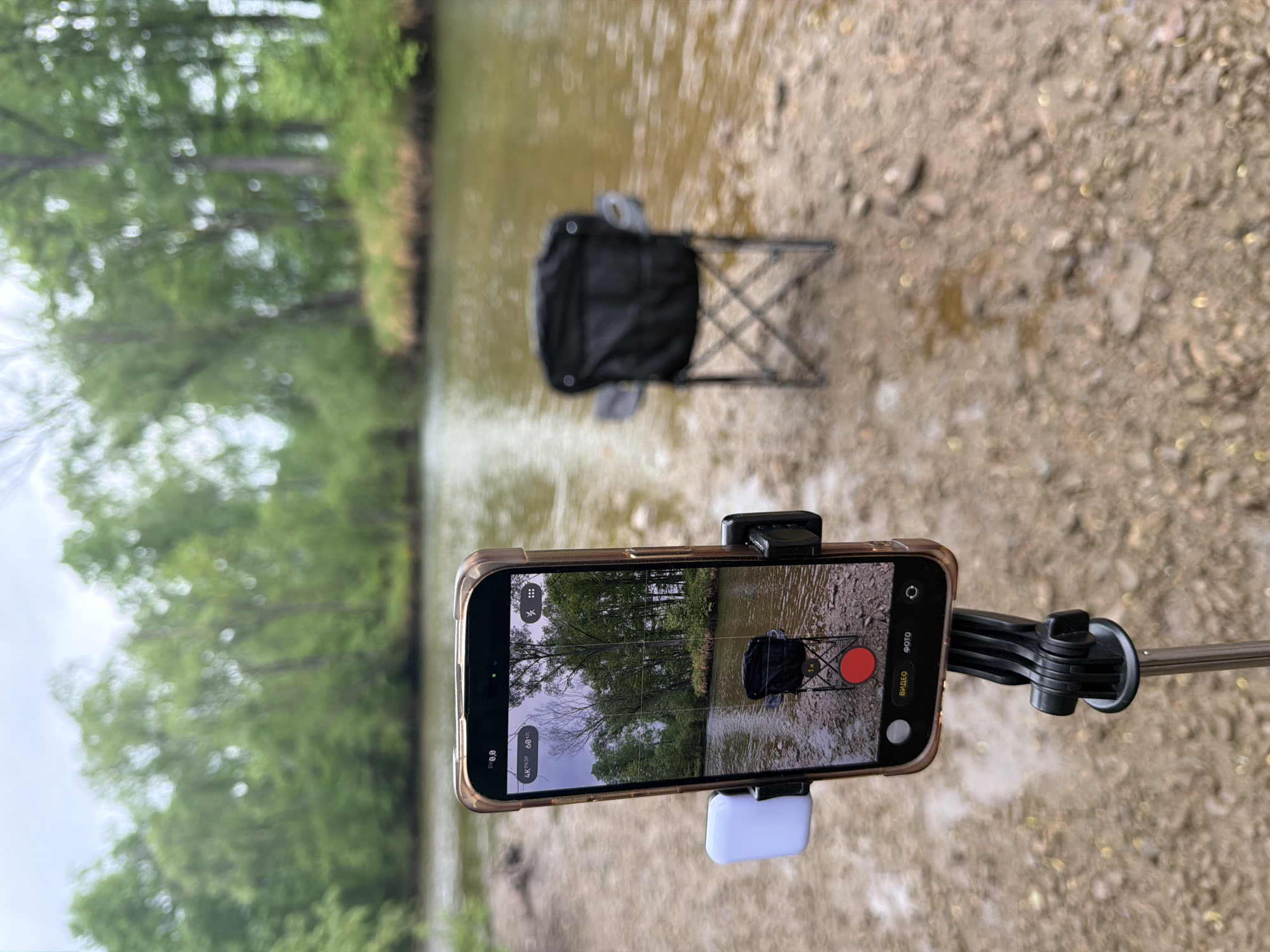 The scene is an outdoor environment near a river or stream, likely in a rural setting. A person is taking a photo or video with their smartphone mounted on a selfie stick. The phone is focused on a black folding chair placed in the shallow water of the stream. The water is murky or slightly colored, and the streambed is rocky. The background includes trees and foliage, suggesting a wooded area. The photograph is taken from a low angle, close to the ground. No people are visible other than the implicit presence of the photographer holding the phone. The location cannot be identified as Trek, Russia, based solely on the visual information provided.