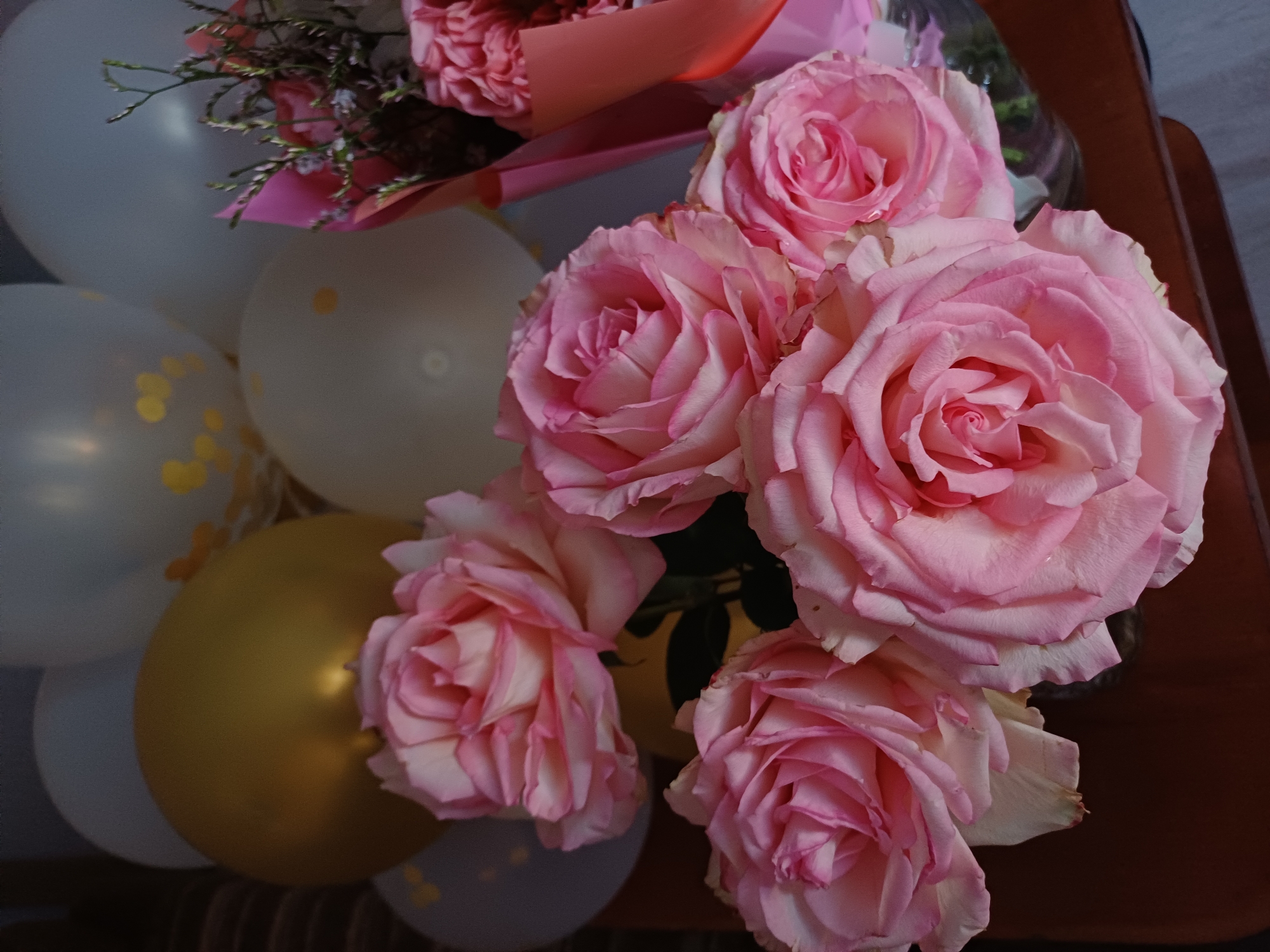 A bouquet of pink roses is displayed in front of a collection of balloons. One balloon is gold, and the others are white with gold speckles. A pink floral wrap is visible to the right of the roses. The scene appears to be indoors, on a wooden surface.