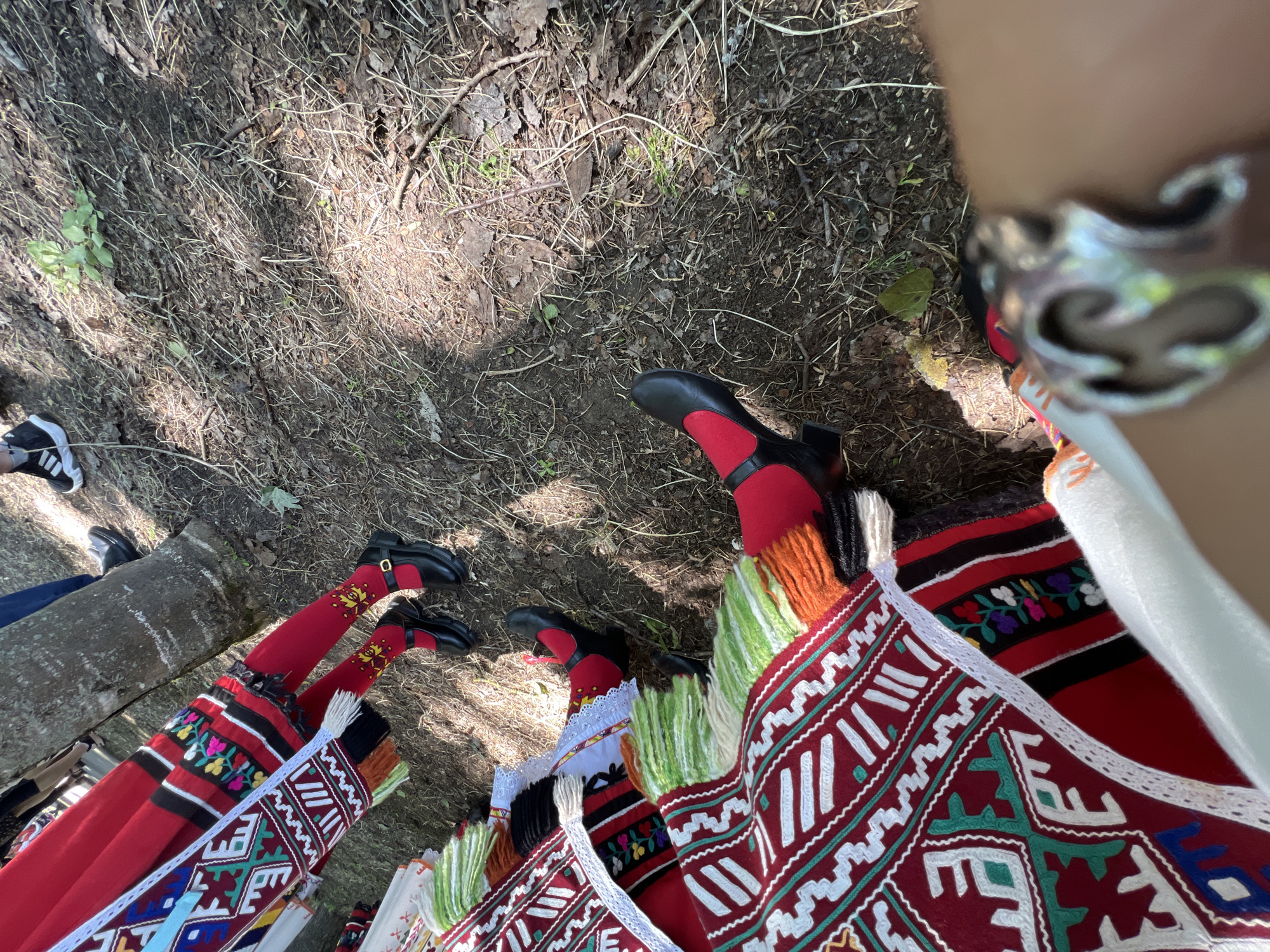 The image captures an outdoor scene in Bolyarovo, Bulgaria, observed from a low perspective. In the immediate foreground, the lower portion of a human arm or wrist, adorned with a prominent silver bracelet featuring an ornate design, is visible.
Two individuals are present, partially visible, dressed in traditional folk costumes. The figure on the left wears a long red garment, possibly a skirt or apron, embellished with horizontal bands of black fabric embroidered with colorful floral and geometric patterns, and trimmed with white lacework. Their legs are covered in red tights, with the ankles featuring distinct yellow and gold stylized leaf embroidery. They wear black Mary Jane-style shoes secured with gold buckles. A white woven strap or sash with multi-colored geometric patterns hangs diagonally from this costume.
The second figure, positioned on the right, is largely obscured, but their lower body displays a vibrant garment. This includes a dark red fabric with white and green geometric embroidery, accompanied by multi-colored fringes in shades of white, green, and orange yarn. One foot is visible, clad in a red tight and a black shoe, similar in style to the other individual's.
The ground surface consists of dry, exposed earth mixed with fallen leaves, small twigs, and scattered patches of green vegetation. Two tree trunks are centrally located in the mid-ground, indicating a wooded or natural environment. In the blurred background, other individuals are faintly visible, including one person wearing blue jeans and black sneakers with white soles. The scene is brightly lit by natural sunlight, casting shadows on the uneven ground.