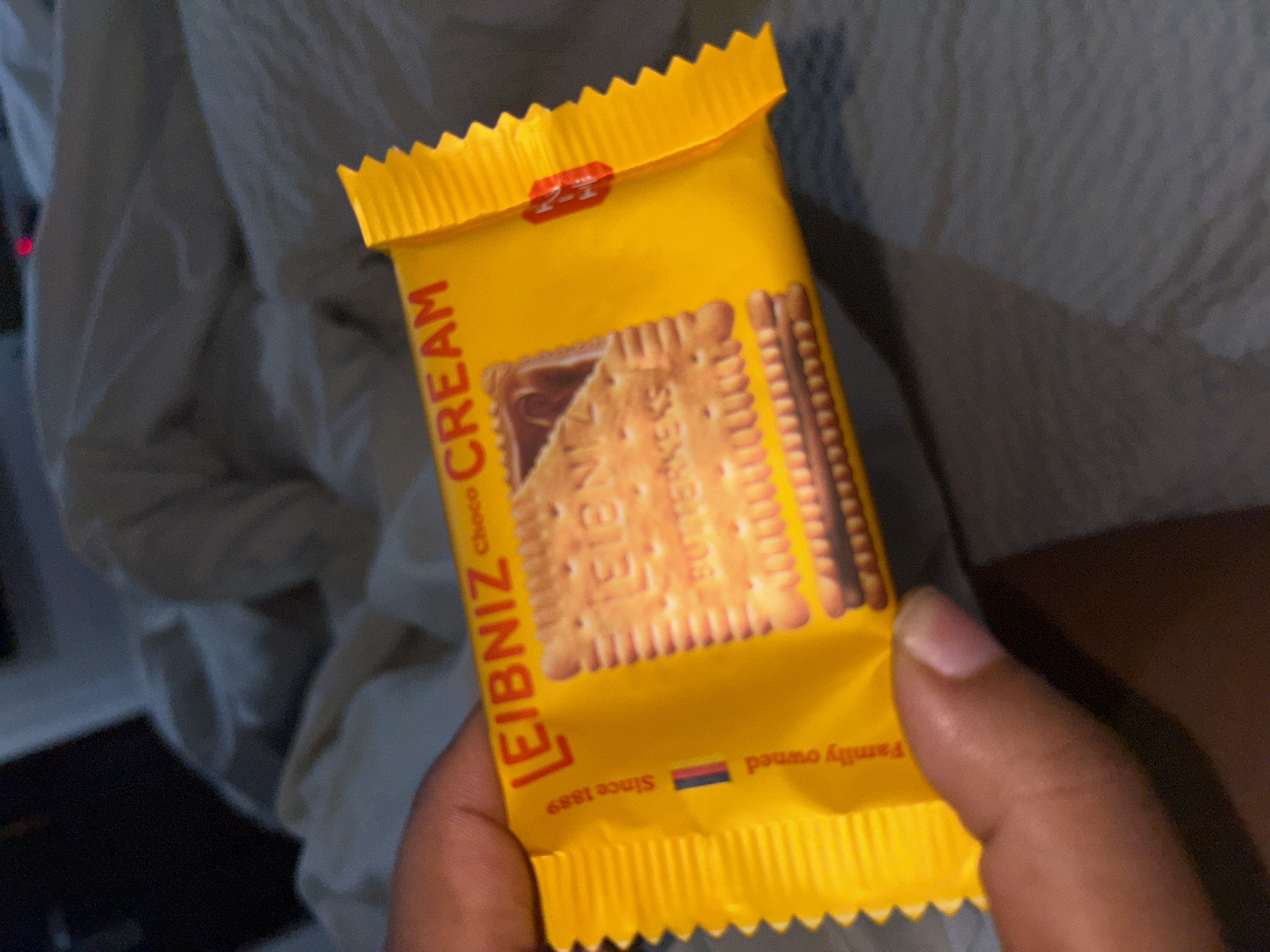 A hand holds a yellow package of "LEIBNIZ Choco Cream" biscuits. The packaging shows an image of the biscuit. The text "Since 1889" and "Family owned" are also visible. The background is slightly blurred and shows what appears to be a white or light-colored cloth. There is no indication of Port Harcourt, Nigeria in the image.