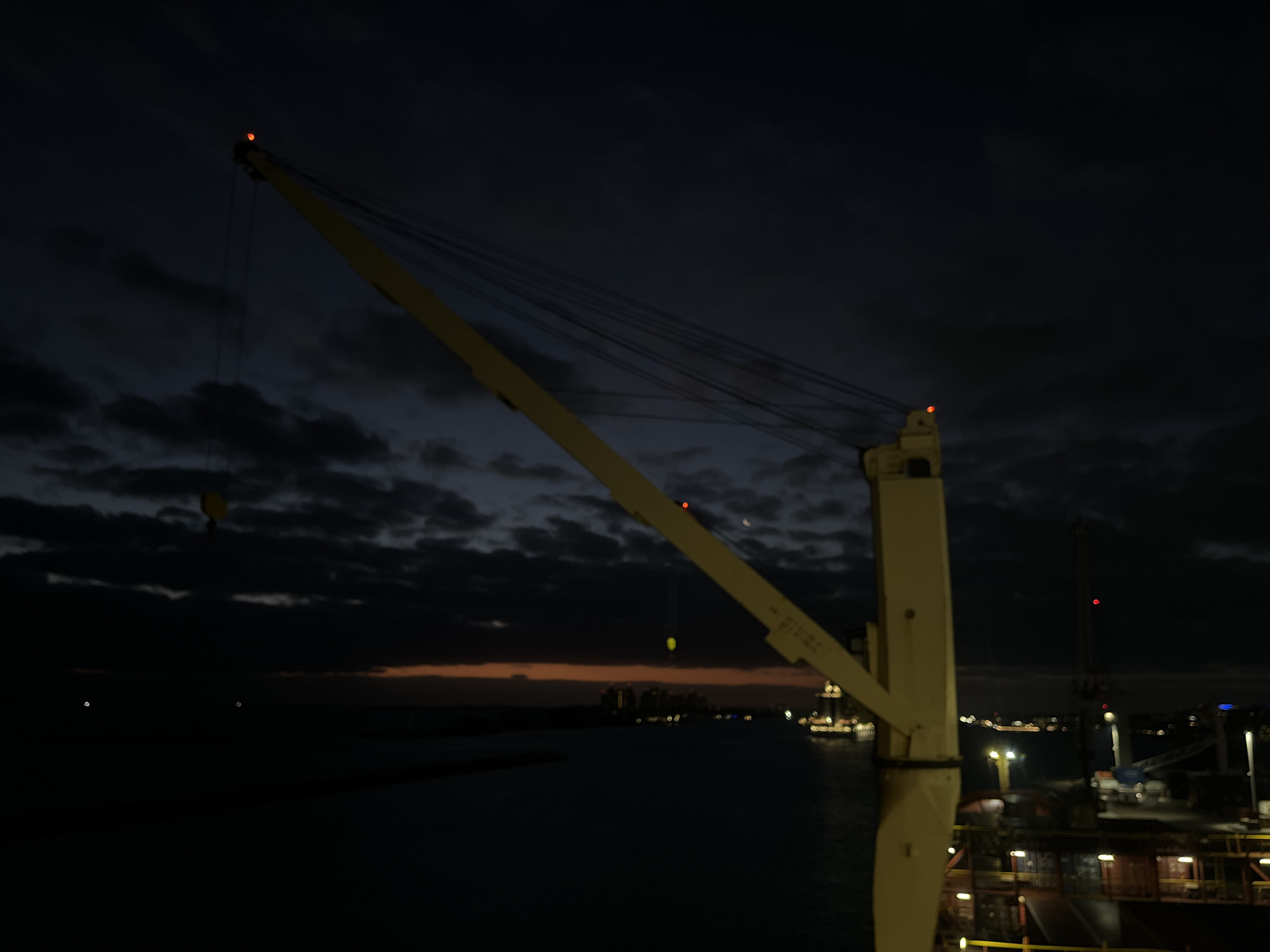 A nighttime scene shows a crane on a ship, with other ships and buildings visible in the background. The sky is dark with clouds, and the water is dark and reflective. The crane is yellow and has a hook hanging from it. There are red lights on the top of the crane and another crane visible in the background.