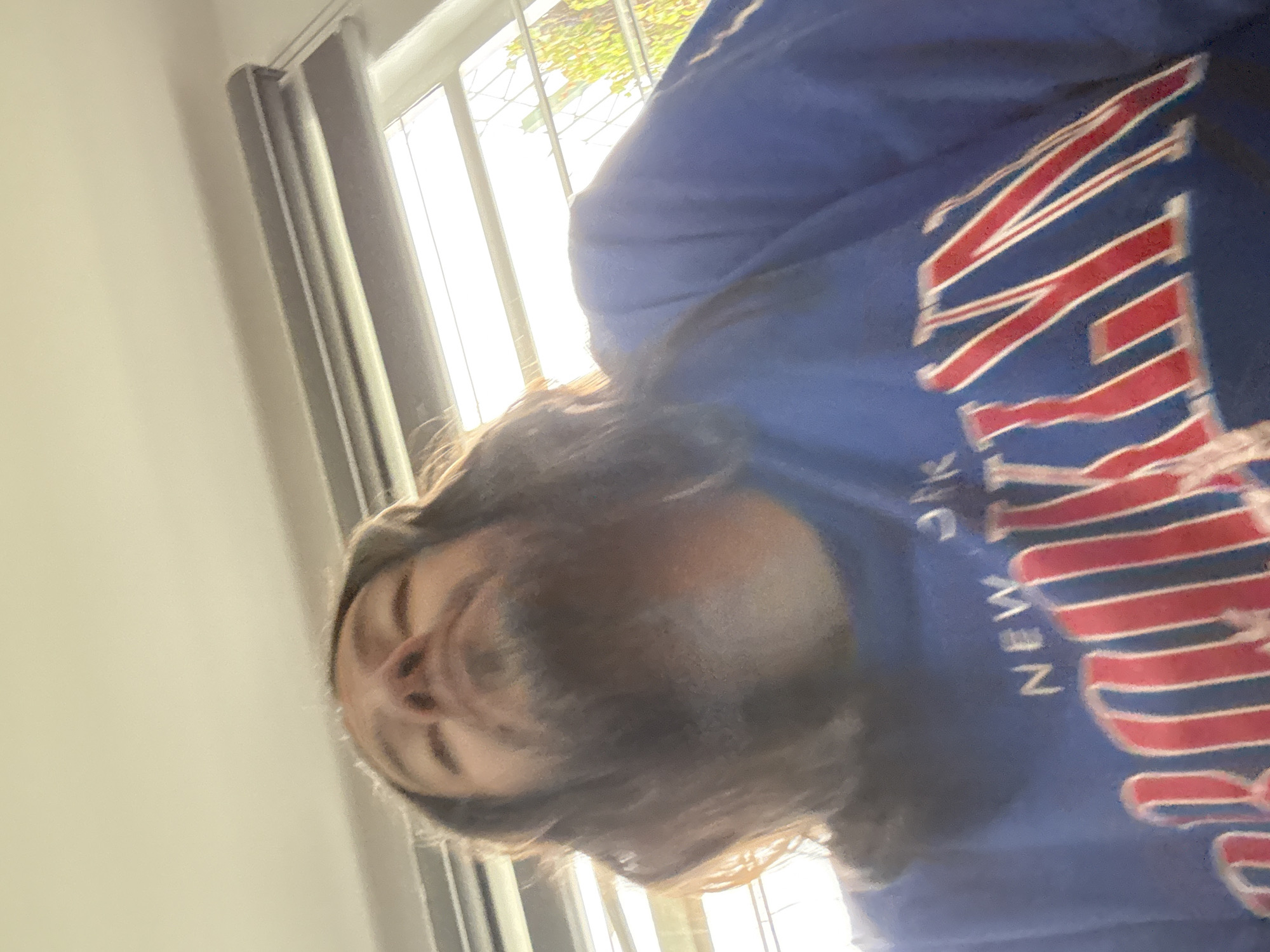 A person is wearing a blue shirt with "New York" written in red and white. The person is inside a building, likely near a window. Outside the window, some trees and bright sunlight are visible. A gray curtain hangs near the window. The image is taken from a low angle, looking up at the person. The specific location in Puebla, Mexico, cannot be determined from the given image.