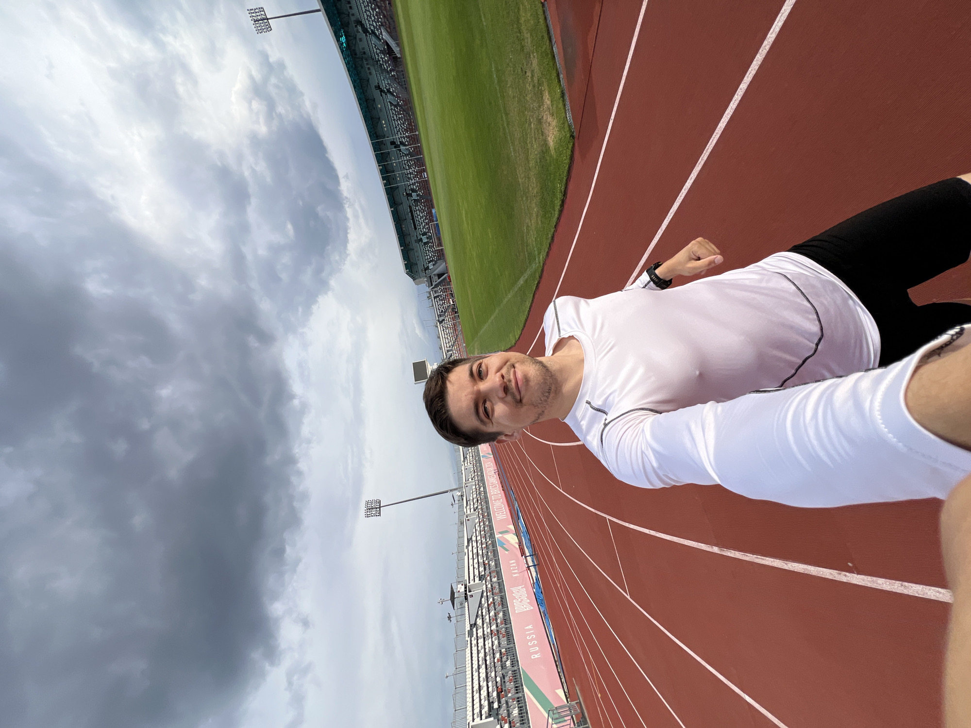 A selfie shows a man on a running track, presumably in the city of Kazan, Russia. The background includes a stadium, track lines, and cloudy sky. The man is wearing a white long-sleeve shirt and black shorts.