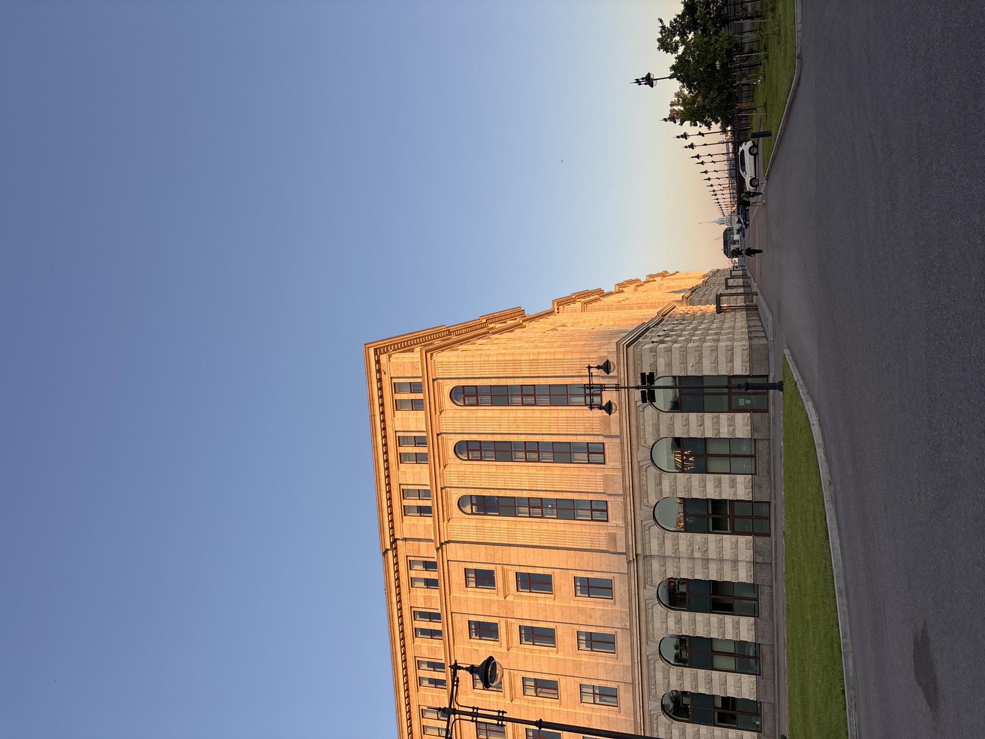 The image shows a wide, paved walkway or road in front of a large, multi-story building with a light stone facade and a cornice. The building has numerous windows of various shapes and sizes, with some reflecting the golden light of a setting sun. The scene is set in Saint Petersburg, Russia, with the building likely being part of the historic city center or a waterfront area, given the presence of a distant bridge and lampposts lining the walkway. The sky is a clear, light blue, suggesting a pleasant evening. There are a few people walking on the walkway, as well as a white car parked on the side. Green grass borders the walkway and the building. The overall impression is one of serene urban landscape bathed in the warm glow of twilight.