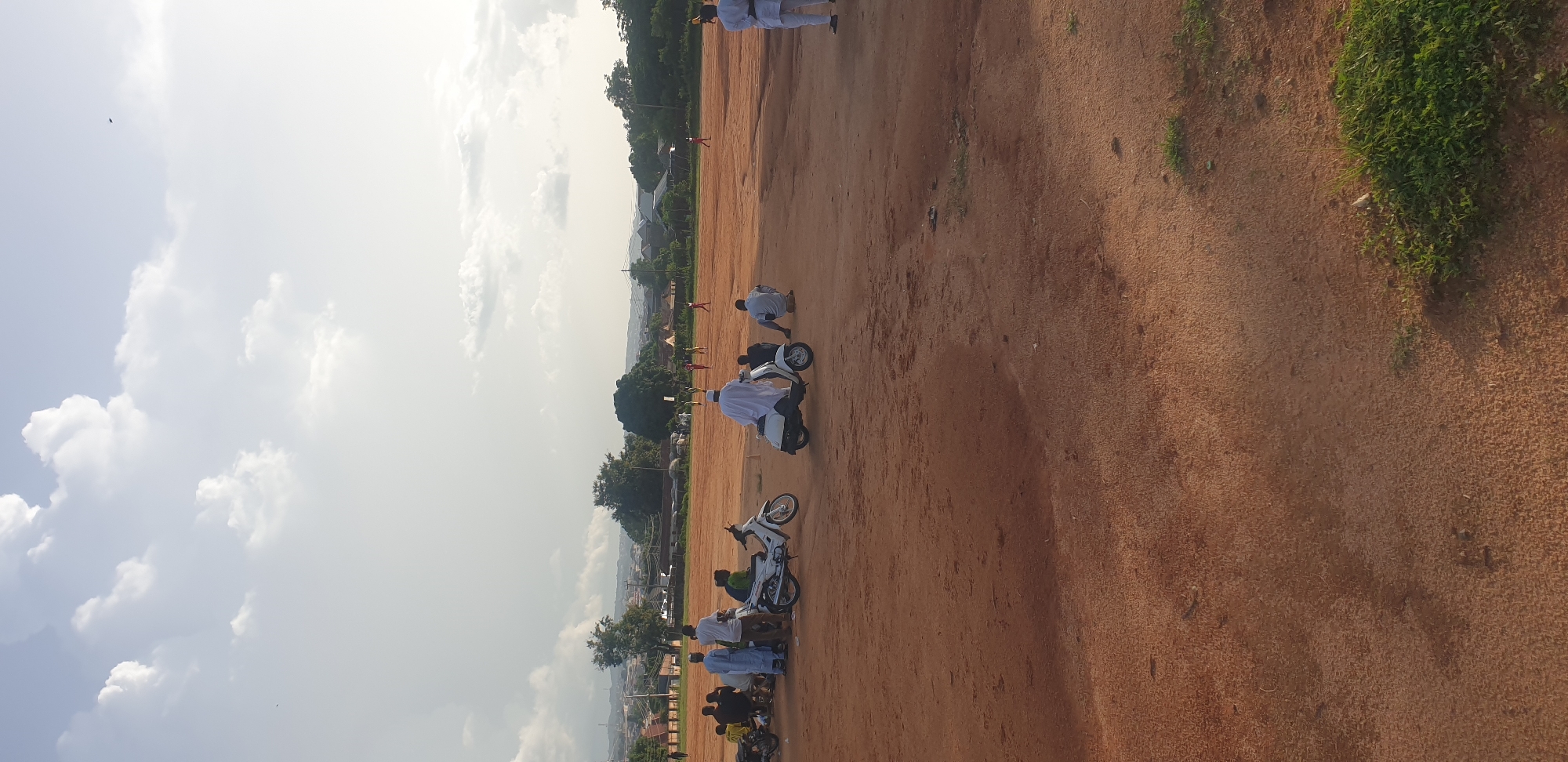 The image depicts an outdoor scene, possibly a sports field or open ground, under a bright sky with scattered clouds. Several people are present, some dressed in white outfits, and a few are on or near motorcycles. In the foreground, there are at least two white motorcycles. One person in a white robe is seated on a motorcycle, while another person in white is crouched beside a different motorcycle. Further back, more individuals are scattered across the reddish-brown ground, with some appearing to be in motion or observing an activity. In the distance, there are trees and buildings, suggesting a populated area. The overall impression is of a gathering or event taking place in an open, possibly dusty, environment. The lighting suggests it is daytime, likely late morning or afternoon, with the sun high in the sky. There is no visible text in the image.