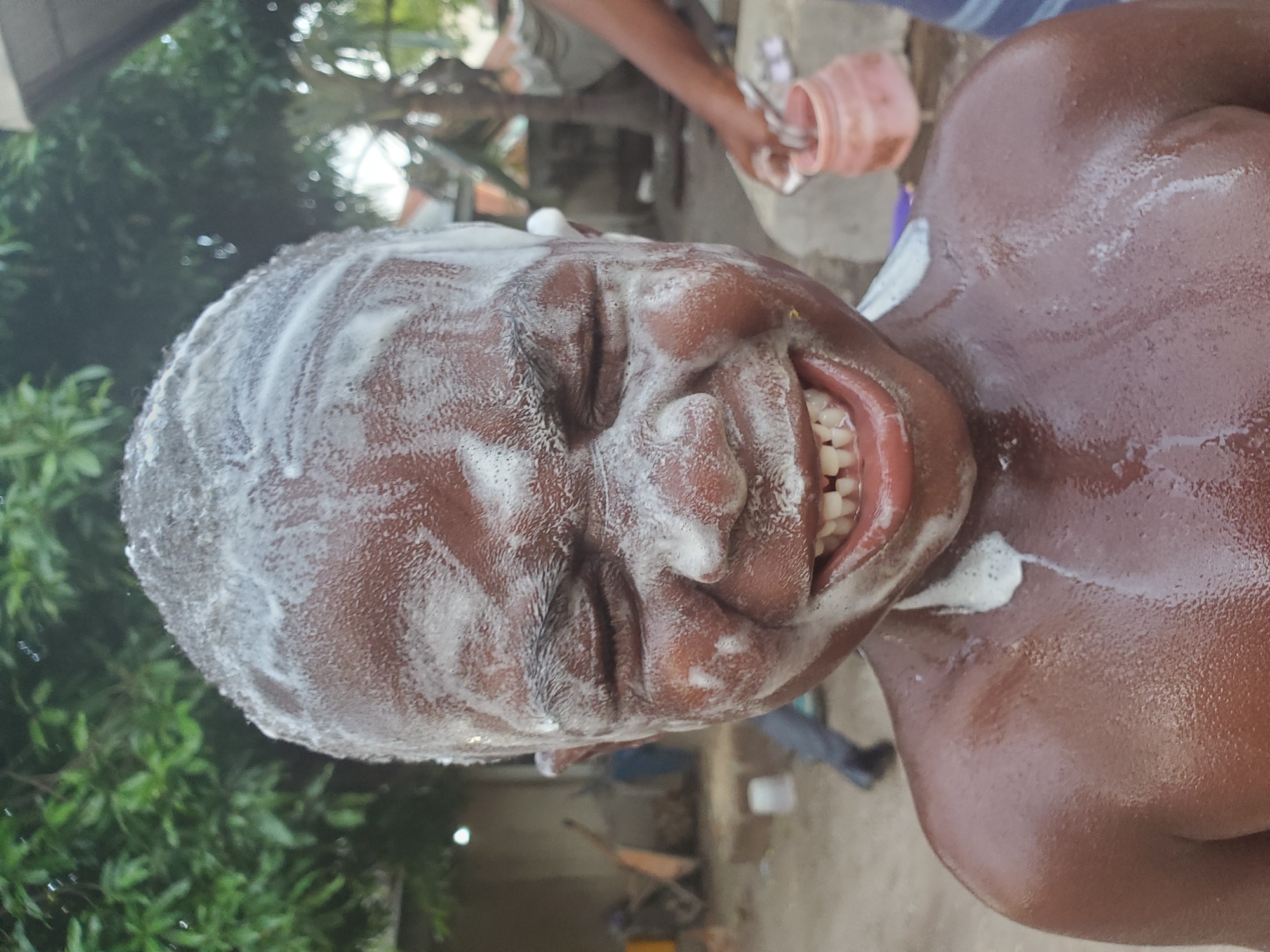 A close-up, high-angle shot depicts a man covered in soap lather from the shoulders up. The man is dark-skinned and has a shaved head. The soap covers his face, including his closed eyes and mouth, which is slightly open, revealing his teeth. The background shows a partially blurred outdoor setting with greenery to the left and a person pouring liquid from a pink container on the right. In the middle ground, there is a building and other objects. The location is Mararaba, Nigeria.