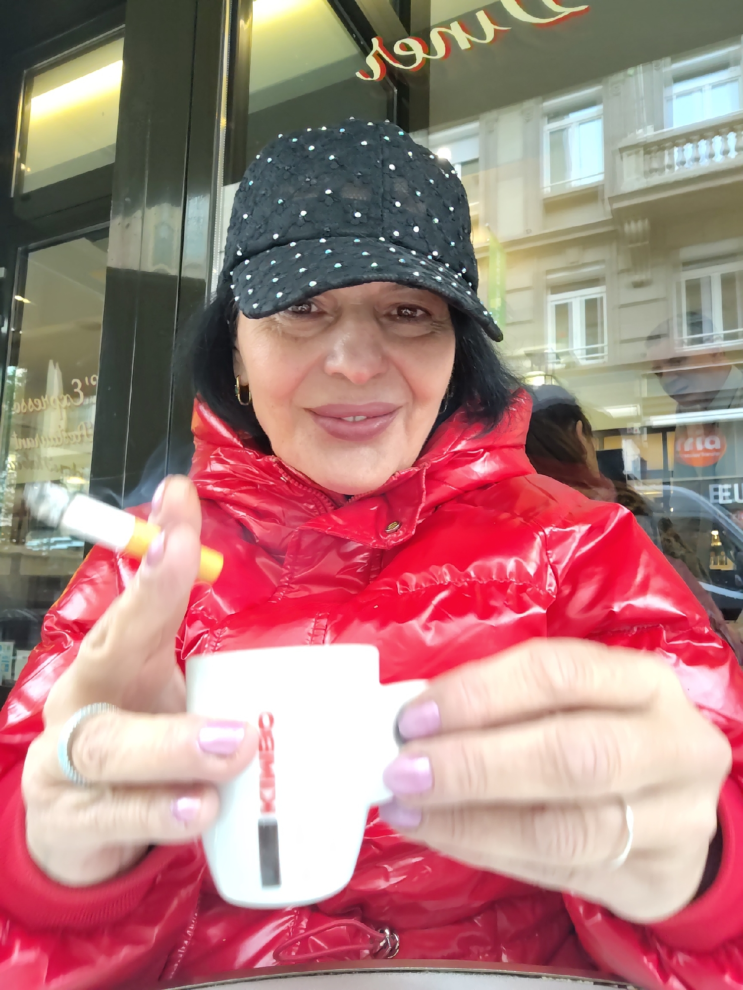 A woman in a glossy red puffer jacket and a black polka-dot hat is sitting at an outdoor café, holding a white coffee mug in her right hand and a lit cigarette in her left. The mug has the word "KIMBO" printed vertically on it. She has dark hair, light pink painted fingernails, and is wearing rings on both hands. Behind her, a glass window reflects a building across the street. Based on the reflection in the window, including the signage "FIA" and architectural style, this is likely a café in Strasbourg, France.
